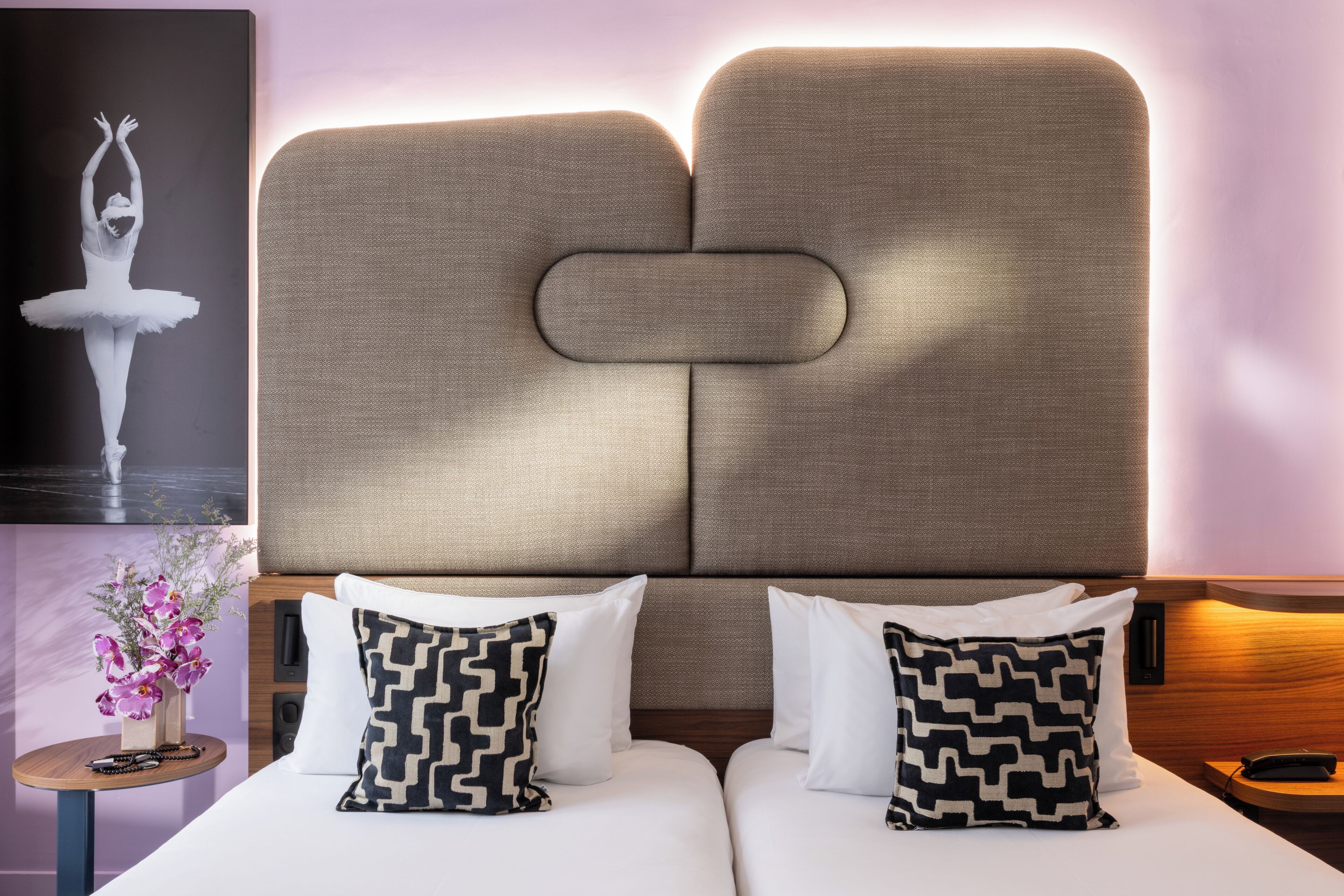 A hotel room with a large bed, two pillows with a geometric pattern, a small side table with a vase of flowers, and a wall-mounted picture of a ballet dancer.