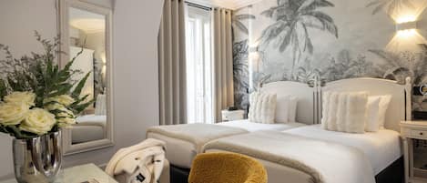 A hotel room with a bed, a chair, a desk with a vase of flowers, a mirror, and a wall with a tropical mural.