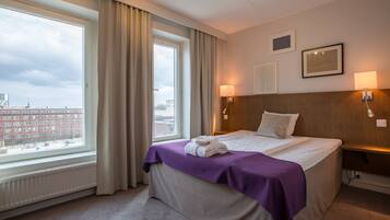 Habitación de hotel con un ventanal grande, cama con colcha morada, dos lámparas de pared y vistas de edificios.