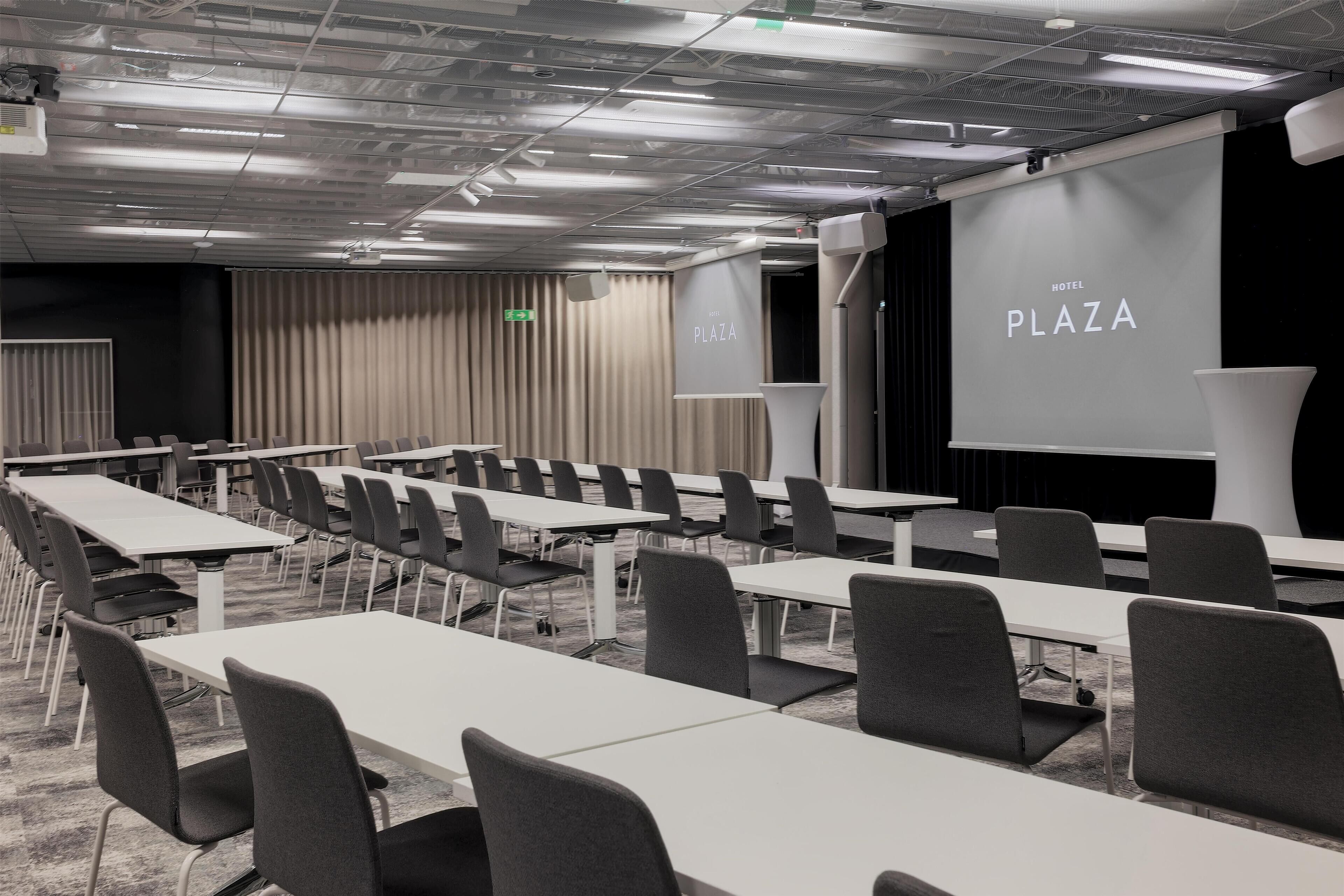 Ein Konferenzraum mit mehreren Tischen und Stühlen, einer Projektionsleinwand mit der Aufschrift "HOTEL PLAZA" und einem Bühnenbereich.