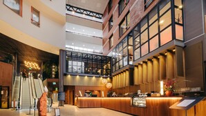 Un hall d’hôtel moderne doté d’un comptoir d’accueil central, d’escaliers mécaniques et d’un plafond haut laissant entrer la lumière naturelle.