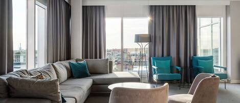 A modern living room with a sectional sofa, two armchairs, a coffee table, and a floor lamp. Large windows with curtains provide a view of the cityscape.