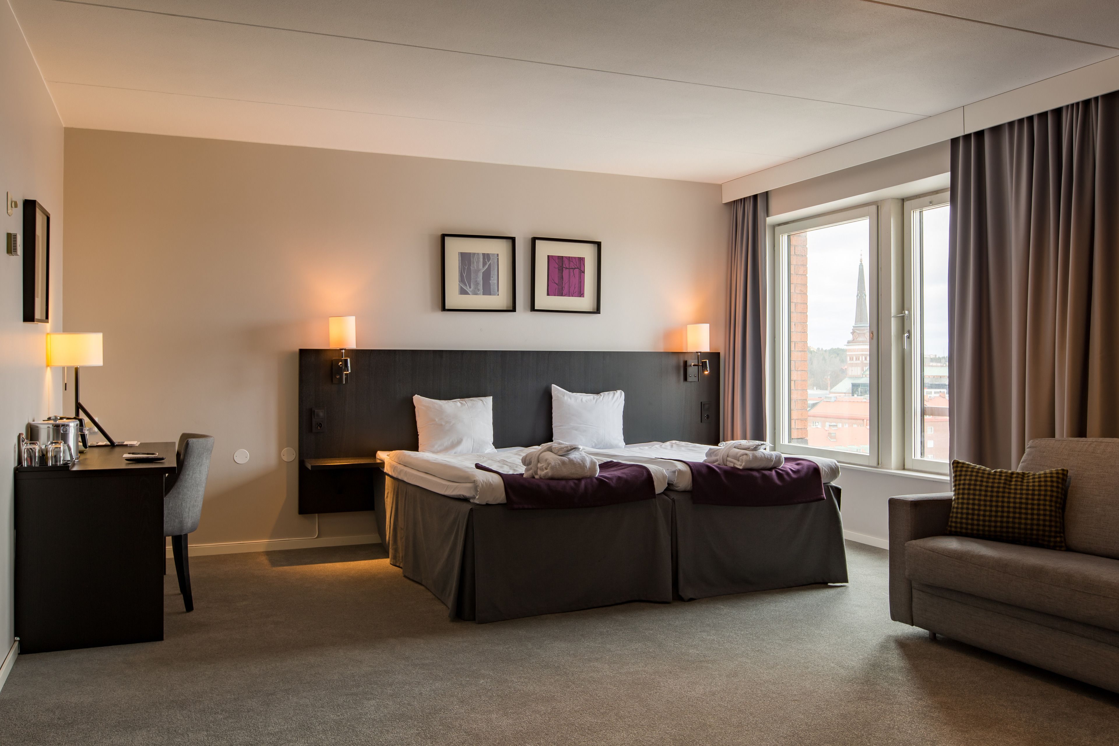 Habitación de hotel con cama, escritorio, silla y sofá. Hay dos cuadros en la pared, una ventana con cortinas y una lámpara en el escritorio.