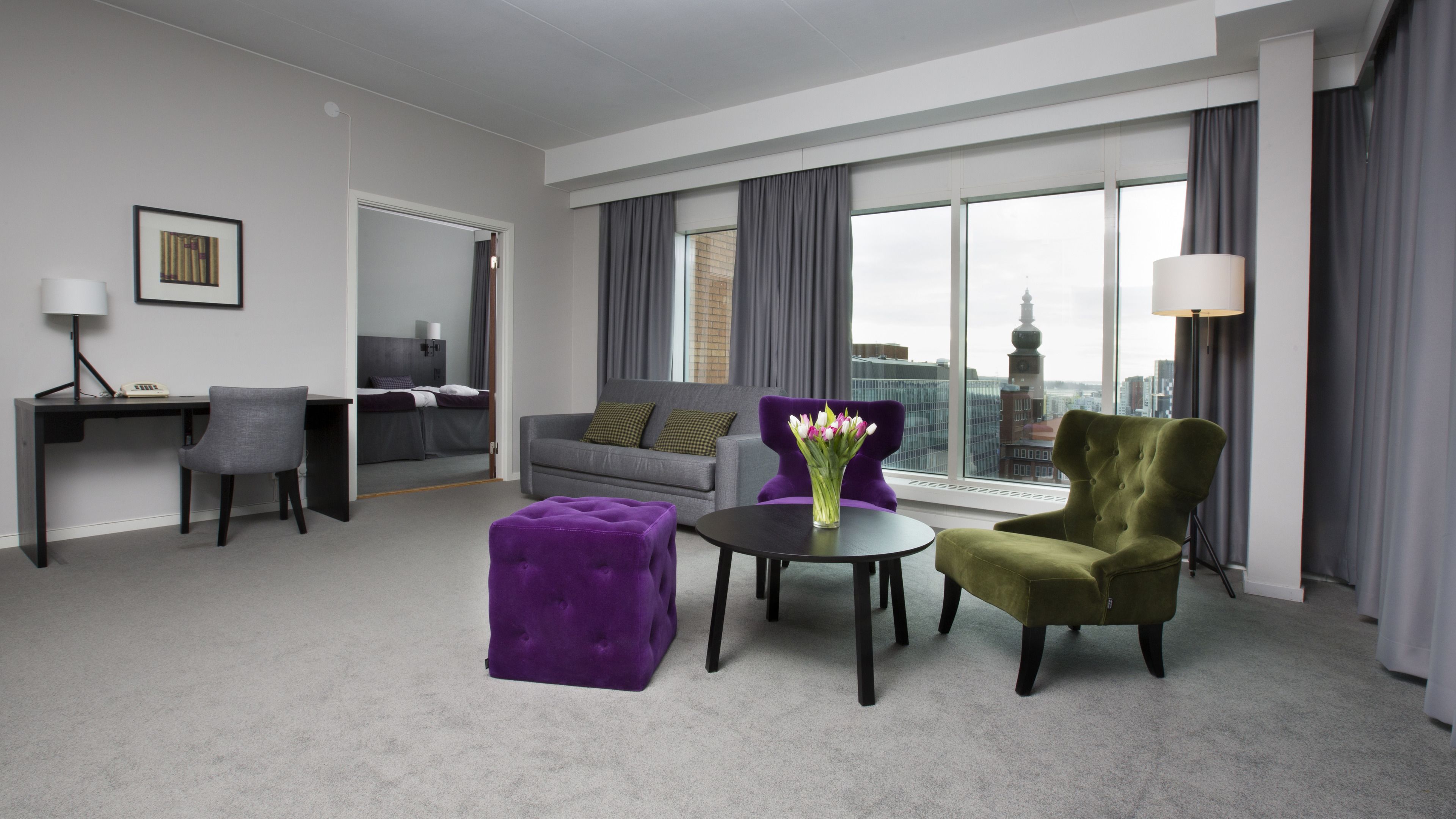 Une chambre d’hôtel moderne avec un canapé gris, un pouf violet, un fauteuil vert et une petite table ronde ornée d’un vase de fleurs. On aperçoit un bureau avec une lampe et une chaise au fond de la pièce.