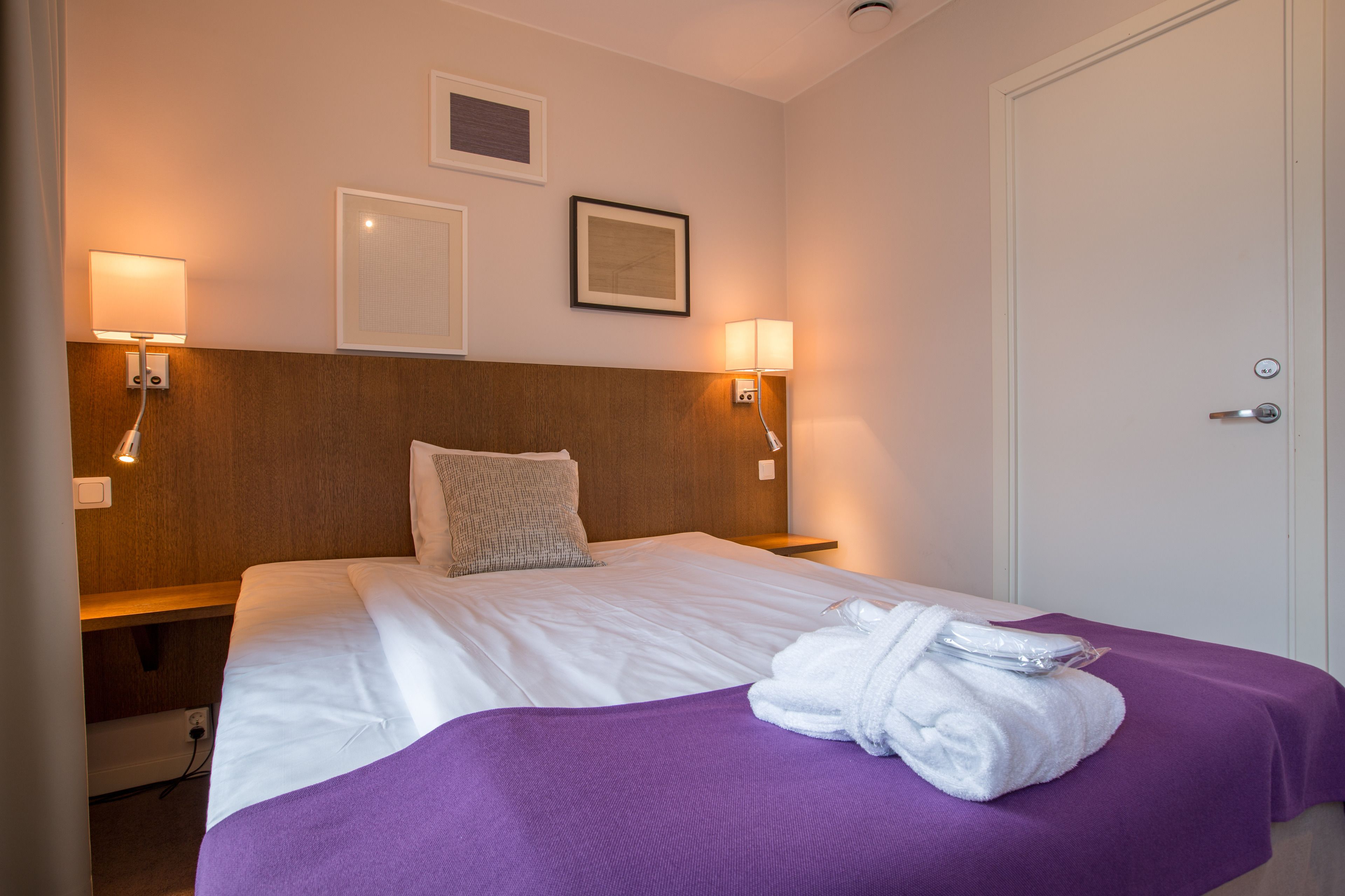 A hotel room with a bed, two wall-mounted lamps, a door, and a small picture on the wall.