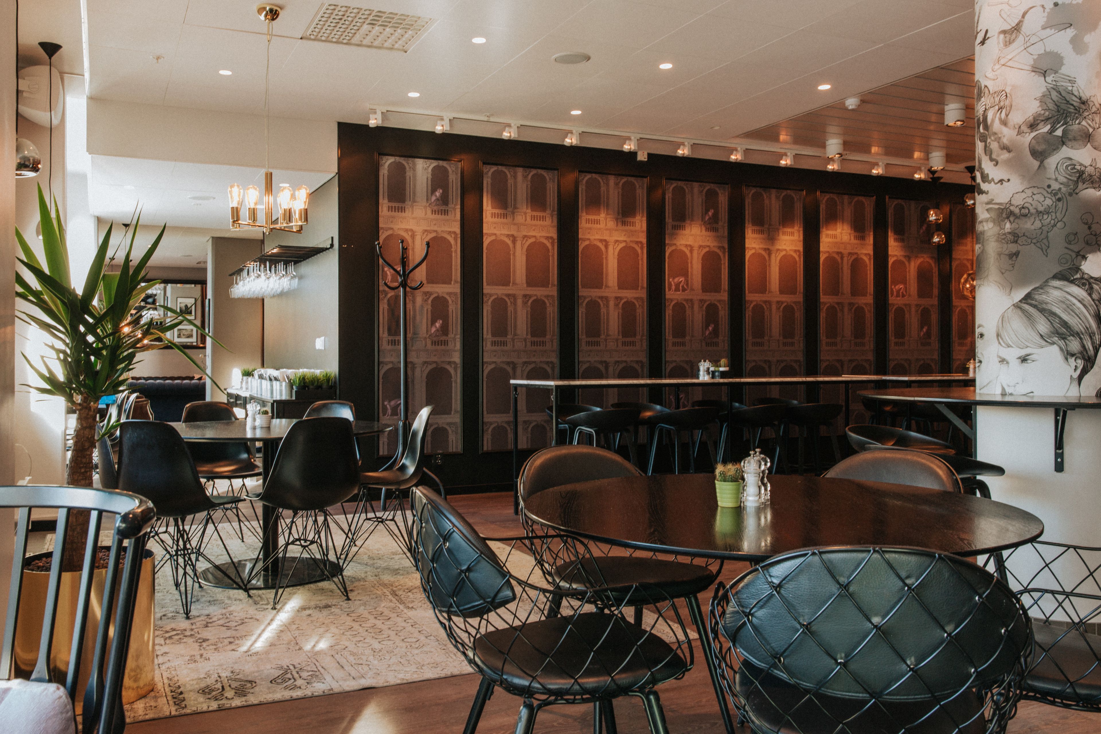 A modern dining area with round tables and black wireframe chairs, a large potted plant, and a decorative wall with a floral pattern.
