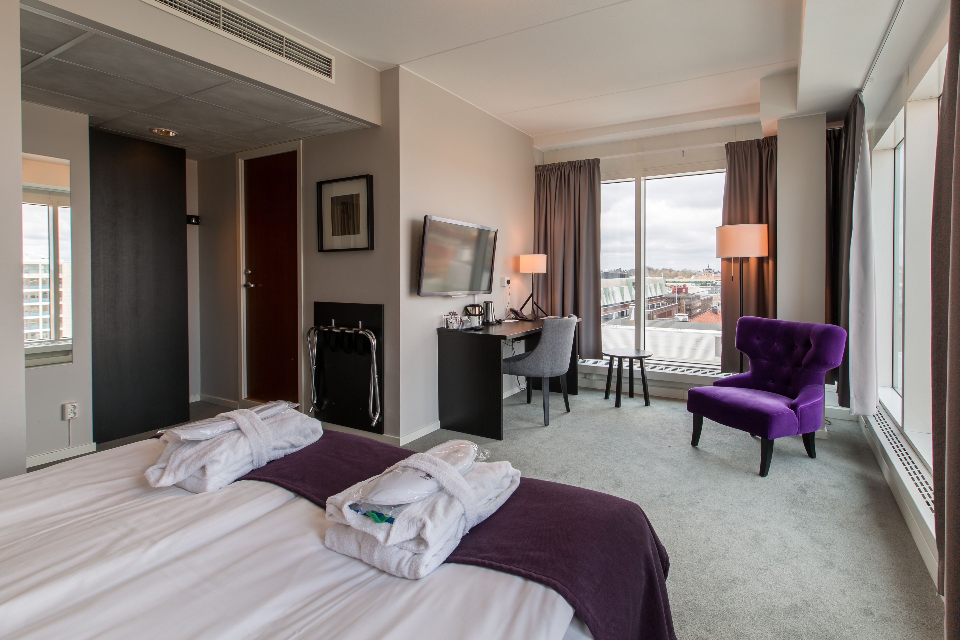 A hotel room with a bed, a desk, a chair, a TV, and a view of the city.