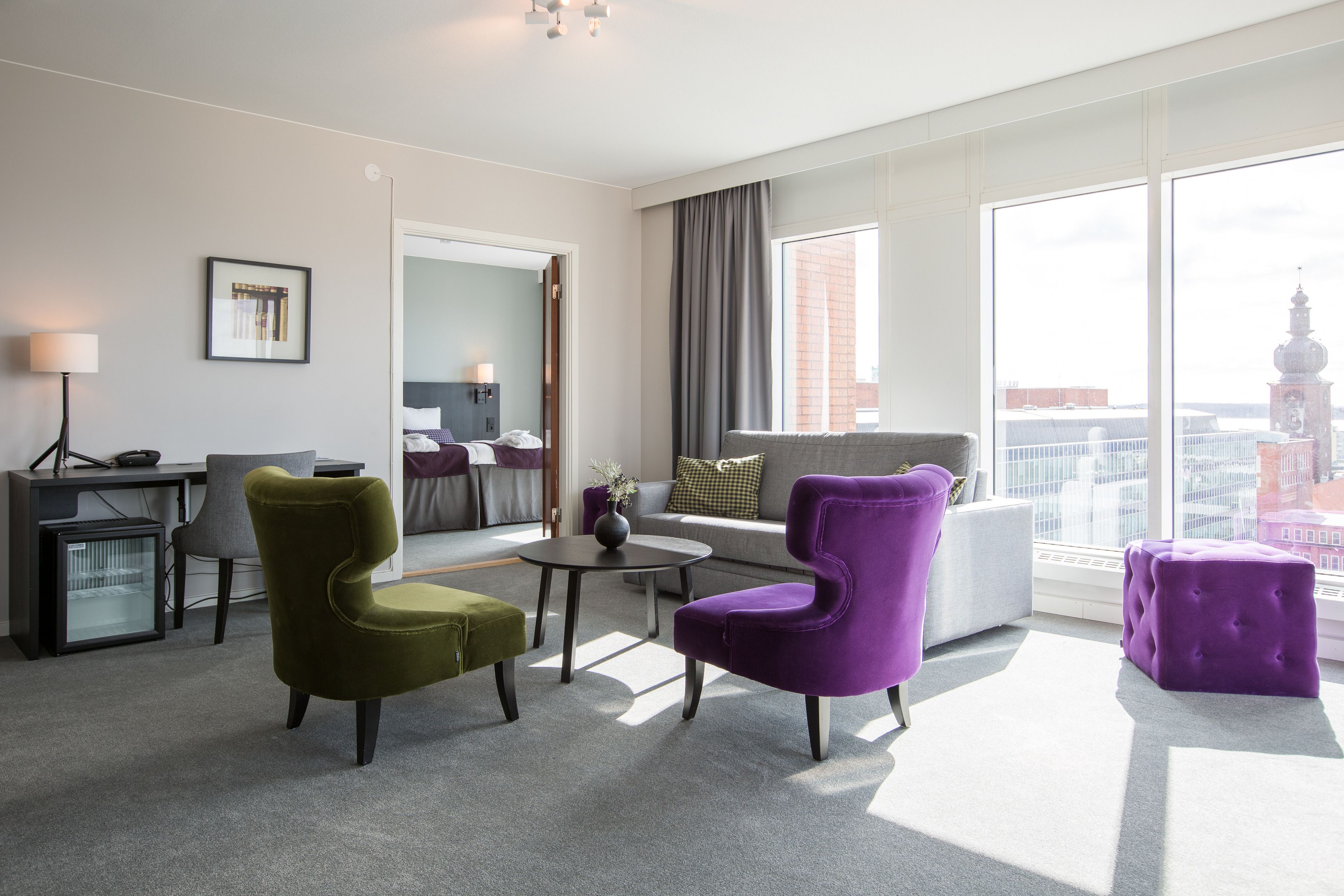 Moderni hotellihuone, jossa on sohva, kaksi nojatuolia, pieni pöytä ja suuri ikkuna, josta avautuu näkymä kaupunkiin.