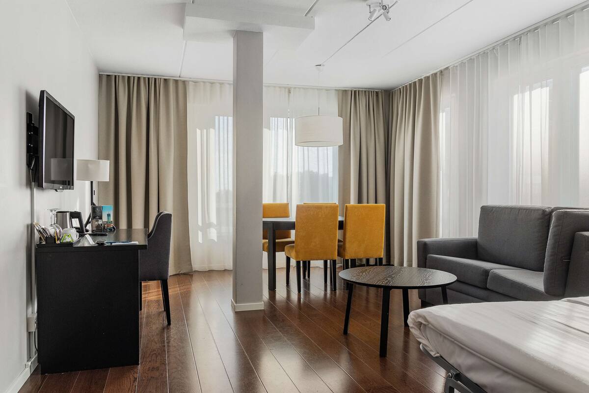 A modern hotel room with a flat-screen TV, a desk with a lamp, a sofa, a dining area with chairs, and a large bed.