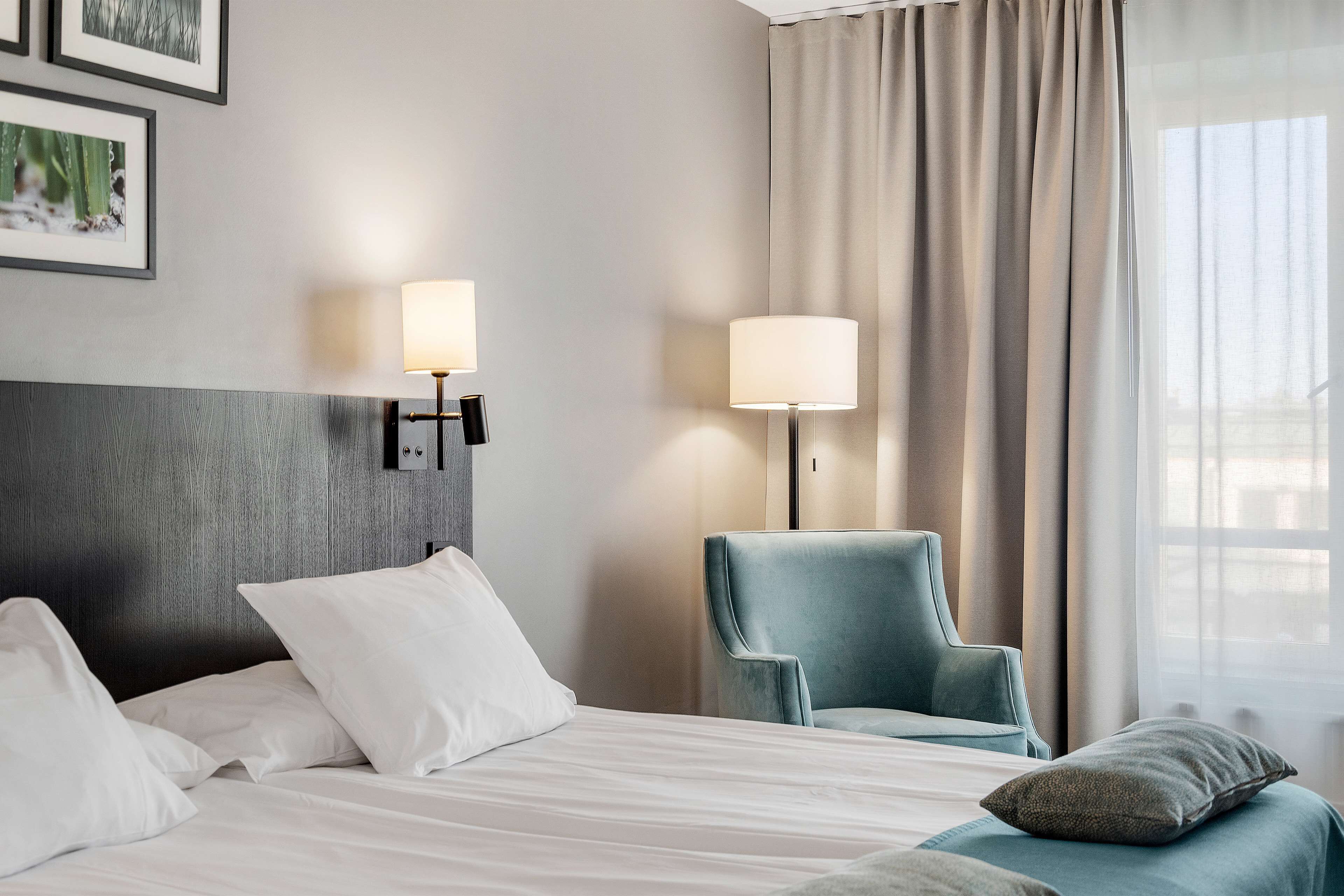 A hotel room with a bed, two wall-mounted lamps, a blue armchair, and a window with curtains.