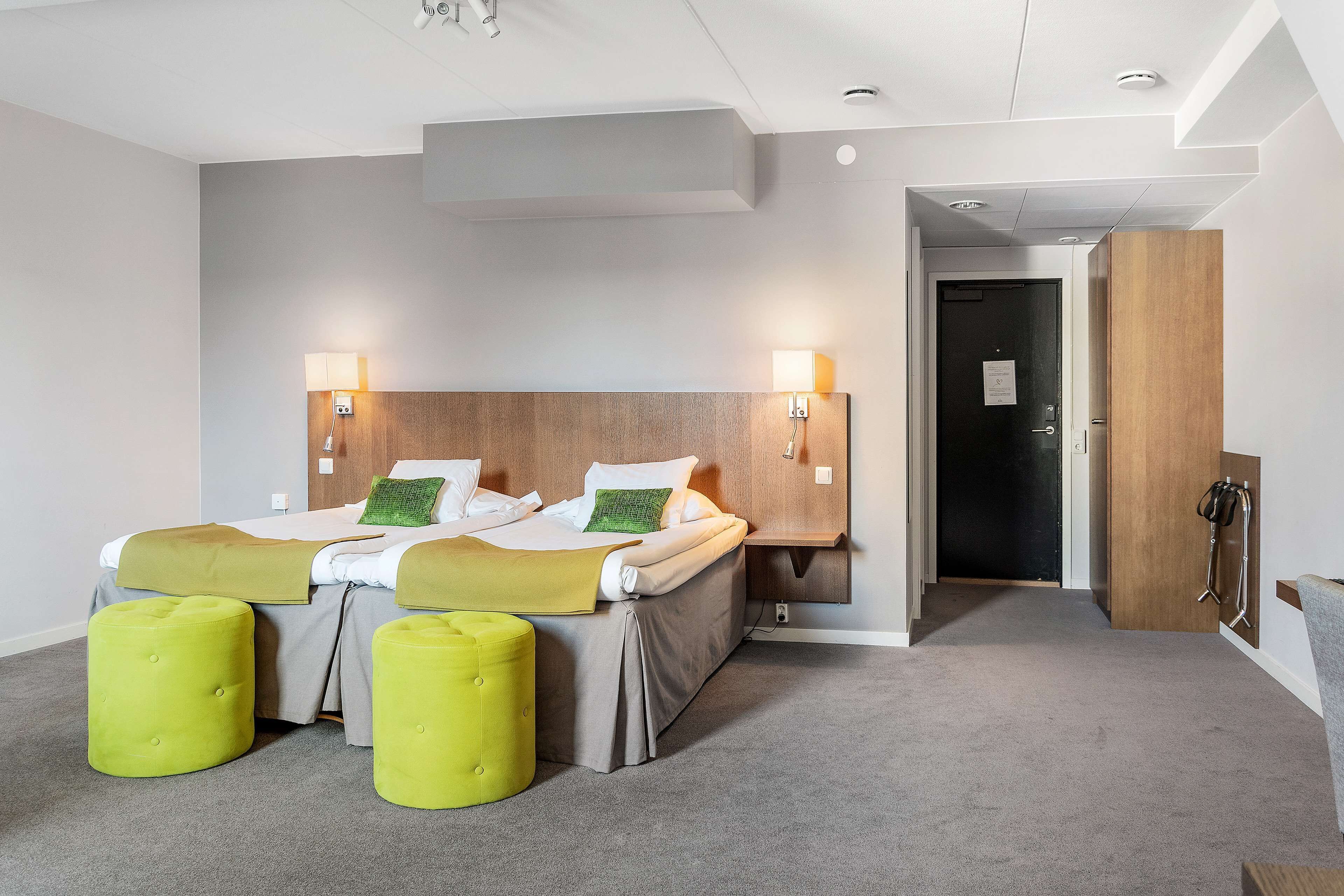 Une chambre d’hôtel avec deux lits, une tête de lit en bois, deux lampes fixées au mur et une moquette grise.