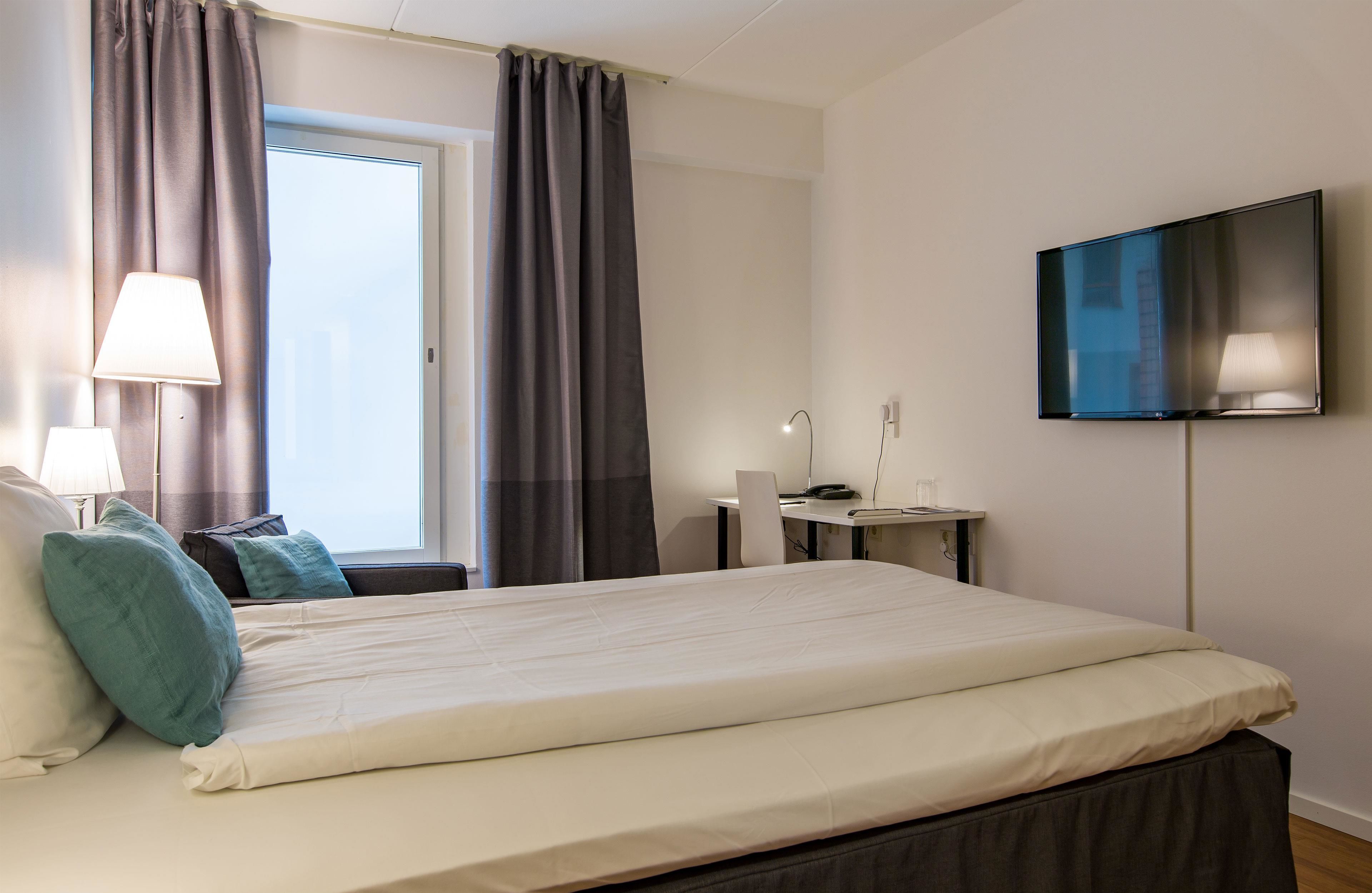 A hotel room with a bed, a desk, a chair, a TV, and a window with curtains.