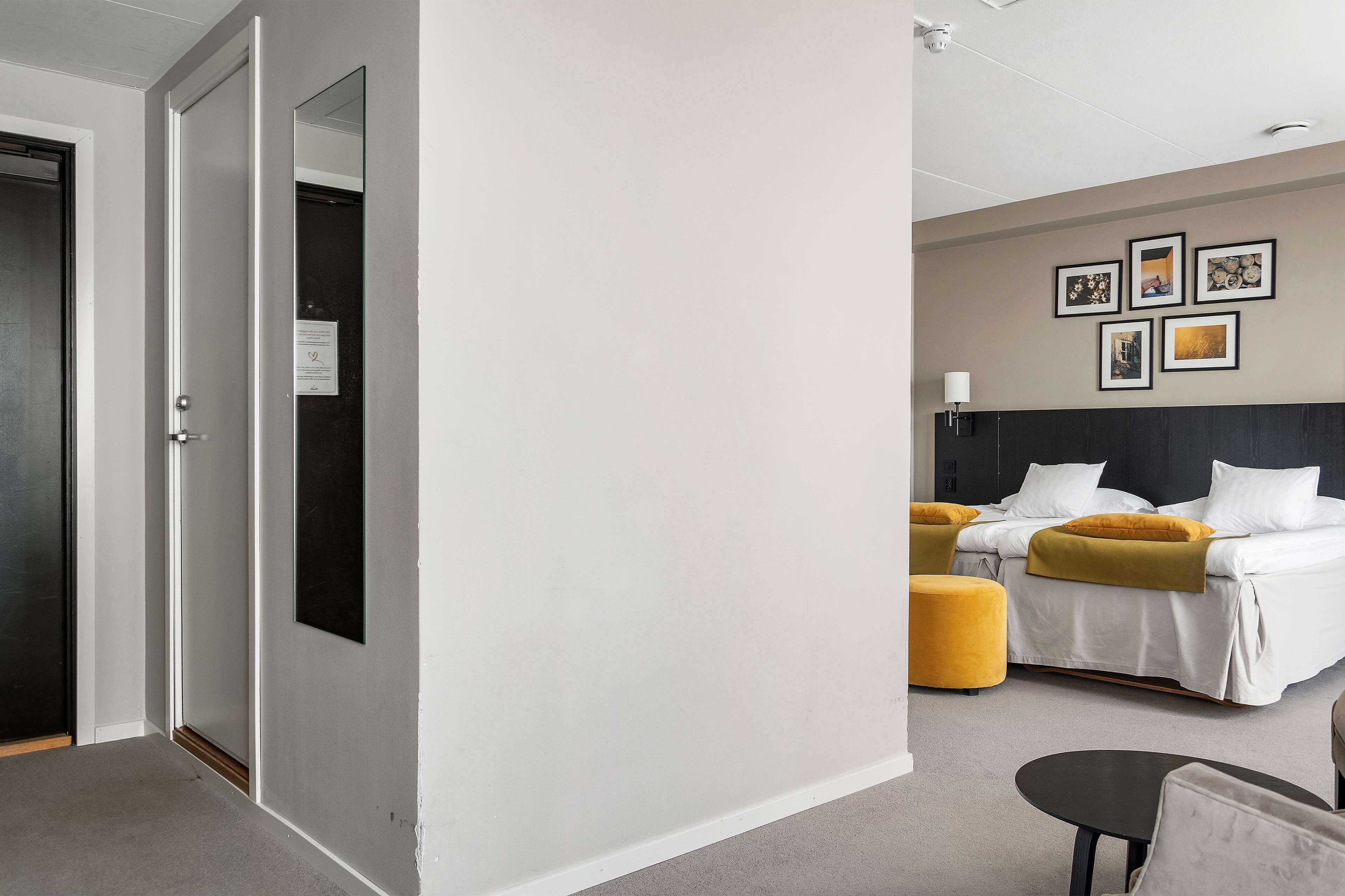 A hotel room with a bed, a bedside table, a chair, and a small table. There are four framed pictures on the wall.