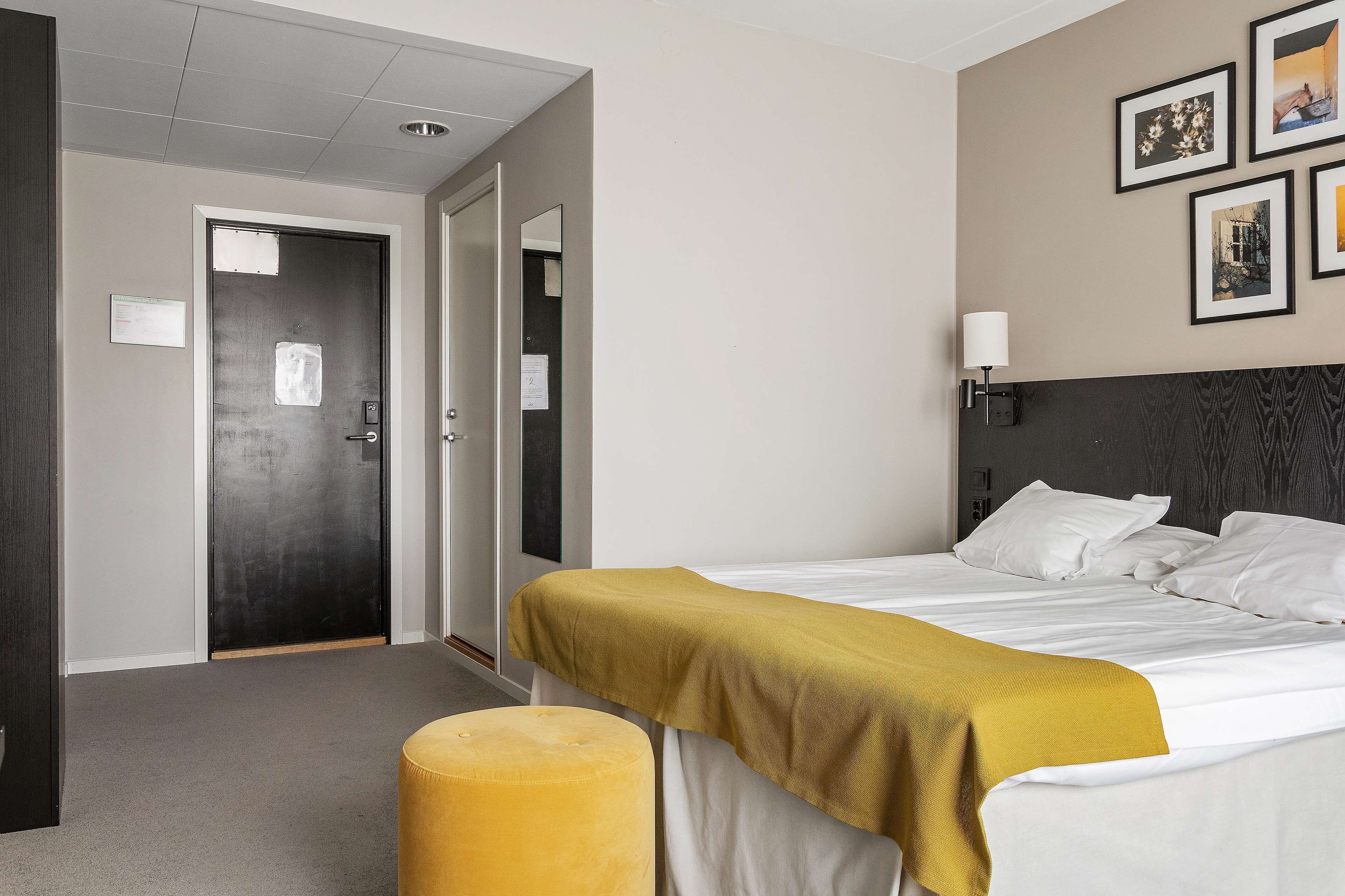 A hotel room with a bed, a yellow ottoman, a wall-mounted lamp, and framed pictures on the wall.