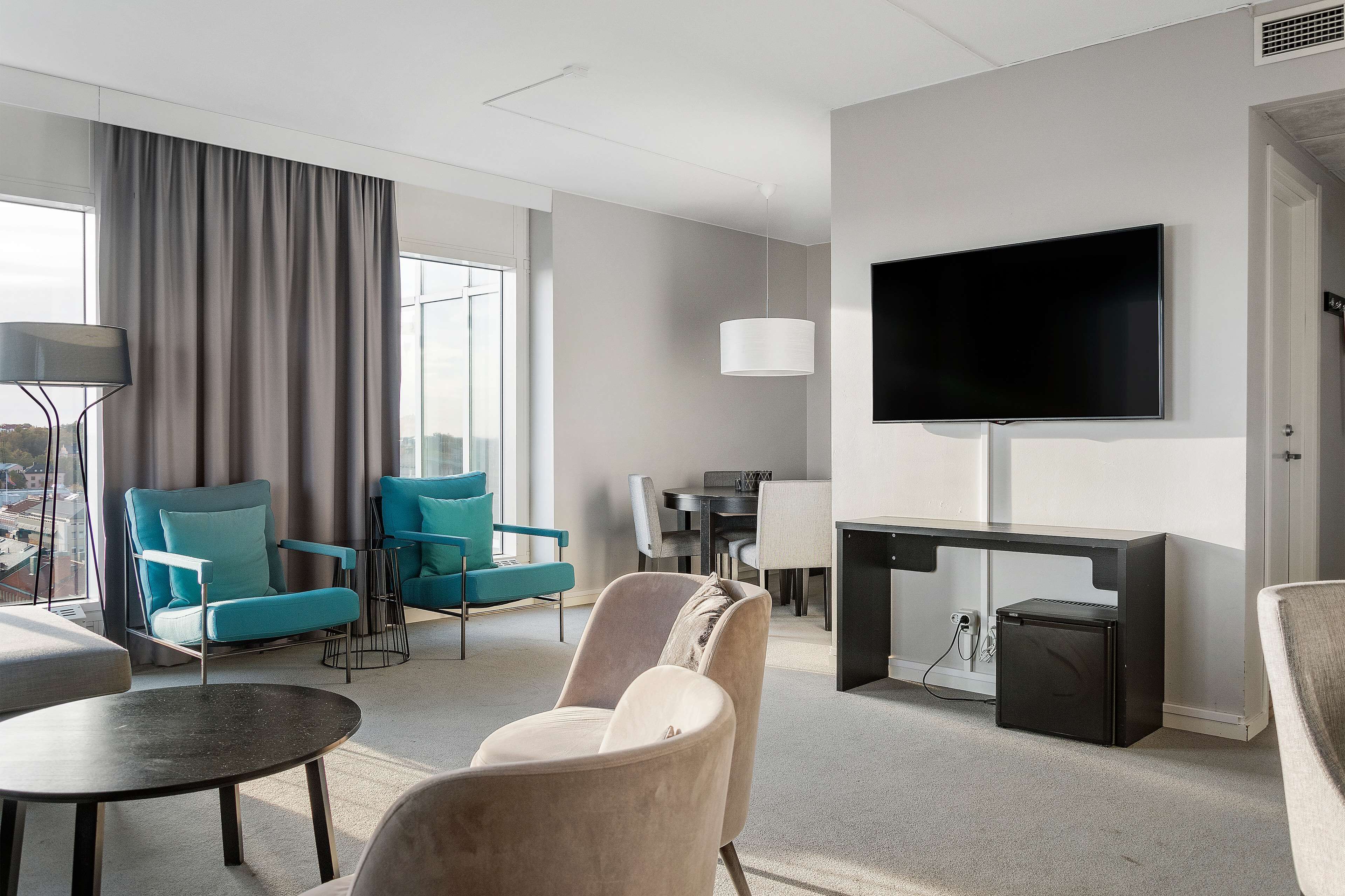 Habitación de hotel moderna con un televisor grande, zona de comedor y asientos con vistas a la ciudad.