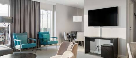 A modern hotel room with a large TV, a dining area, and seating with a view of the city.