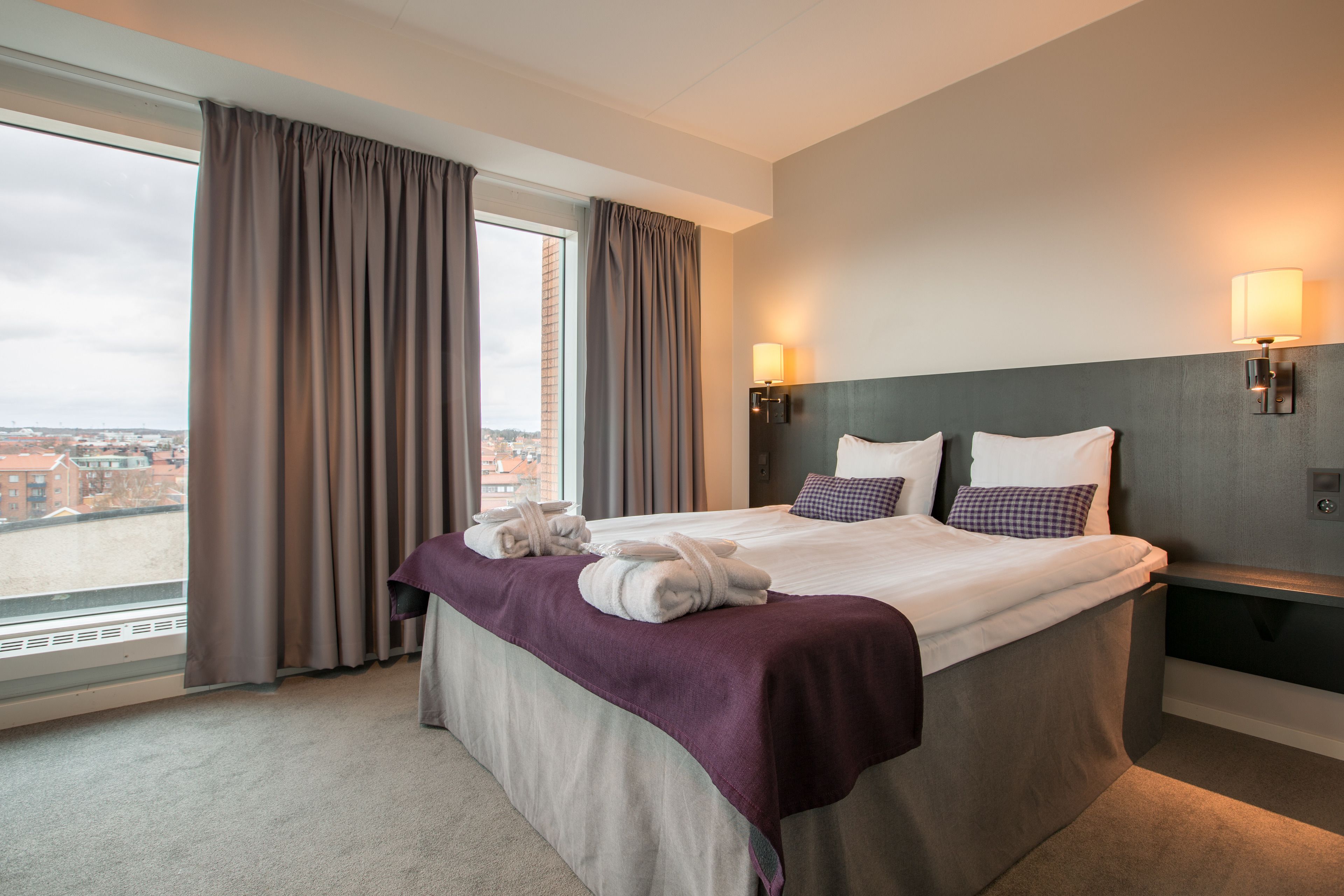A hotel room with a large bed, a view of the city, and two wall-mounted lamps.