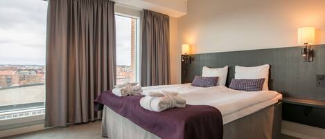 A hotel room with a large bed, a view of the city, and two wall-mounted lamps.