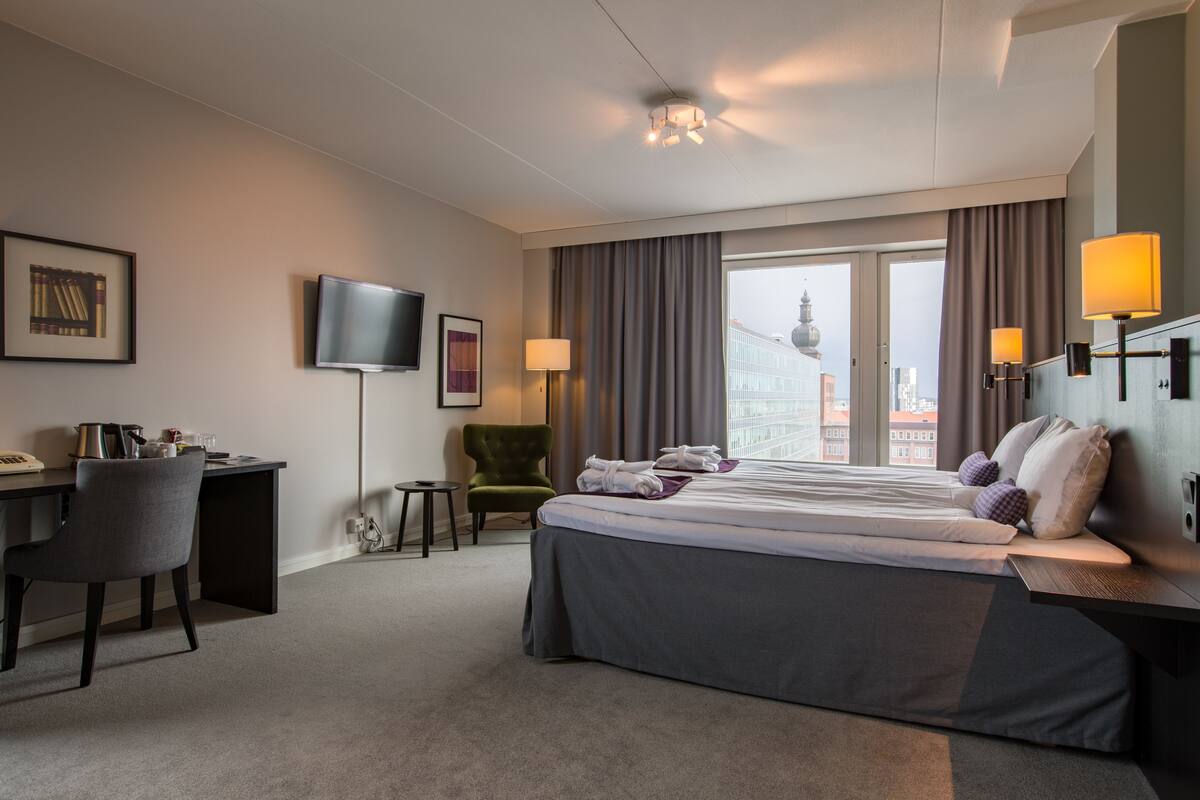 A hotel room with a bed, desk, chair, TV, and large windows with curtains.