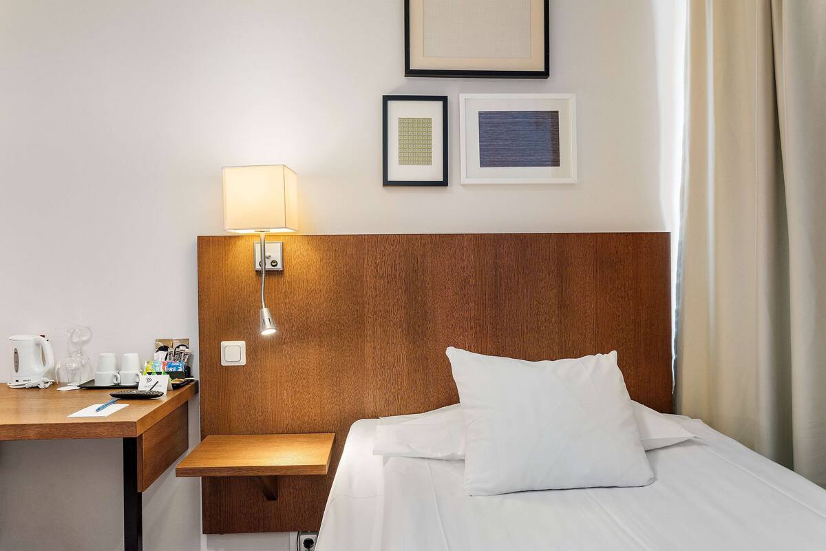 A hotel room with a wooden headboard, a bed with white linens, a bedside table with a lamp, a coffee maker, and a framed picture on the wall.