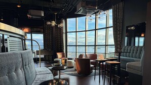 Un salon moderne doté d’un canapé, de poufs et d’un coin bar, avec de grandes fenêtres offrant une vue sur la ville.