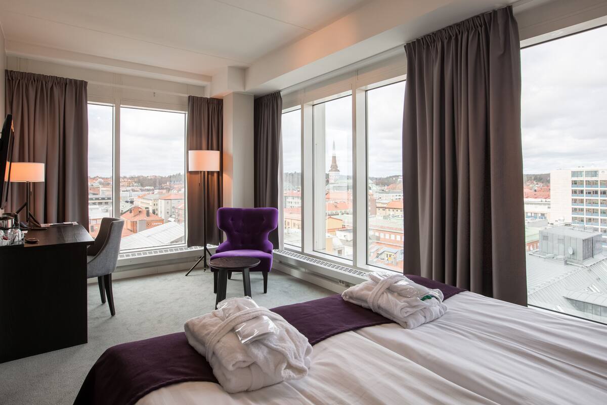 A hotel room with a bed, a desk, a chair, and large windows offering a city view.