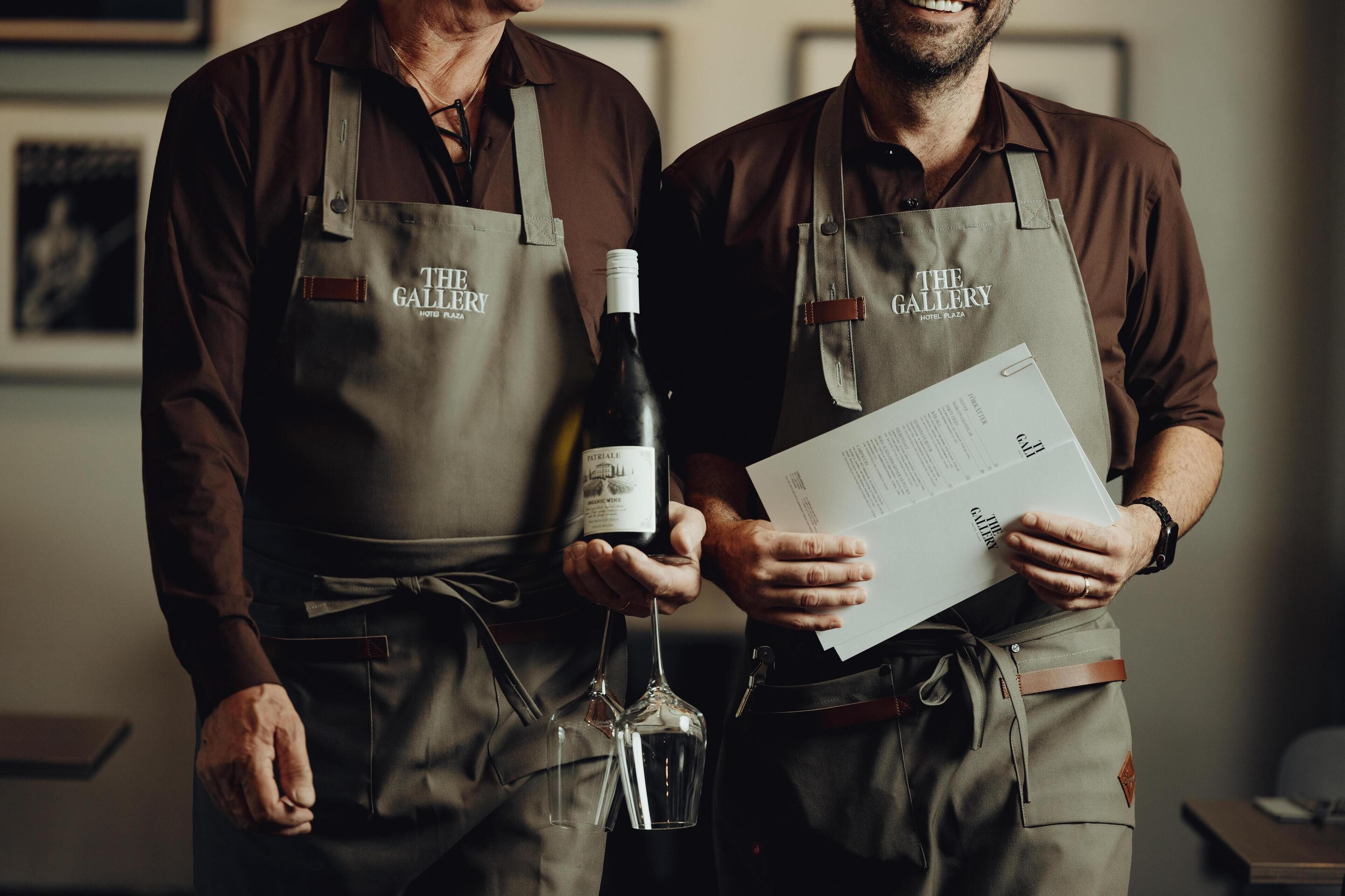 Dos personas con delantales que llevan el logotipo de “The Gallery”, sosteniendo una botella de vino y un menú.