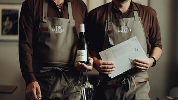 Two individuals wearing aprons with "The Gallery" logo, holding a wine bottle and a menu.