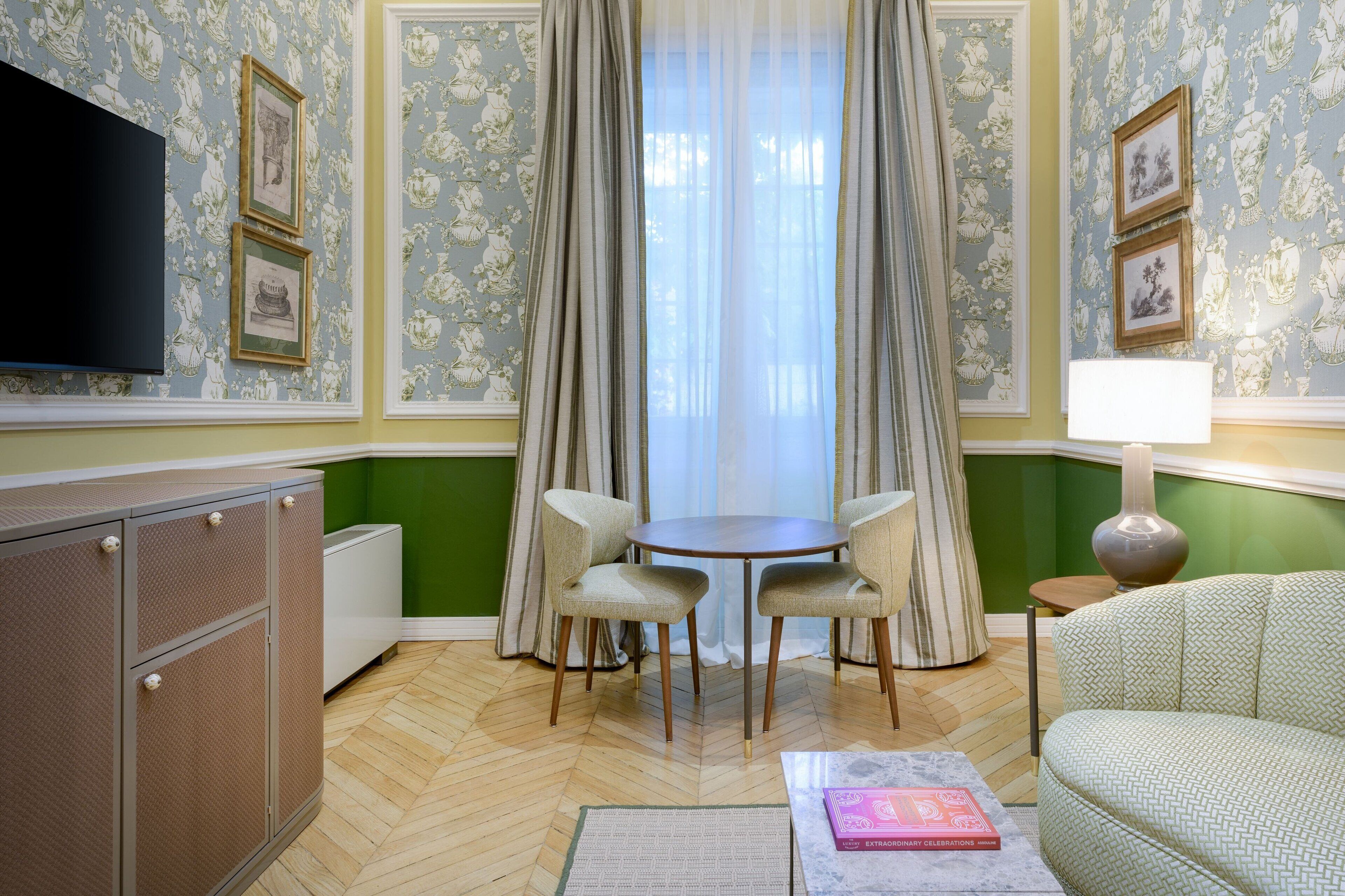 Een kamer met een groene wand, een ronde tafel met vier stoelen, een bank, een televisie die aan de wand is bevestigd, en een lamp op een bijzettafeltje.