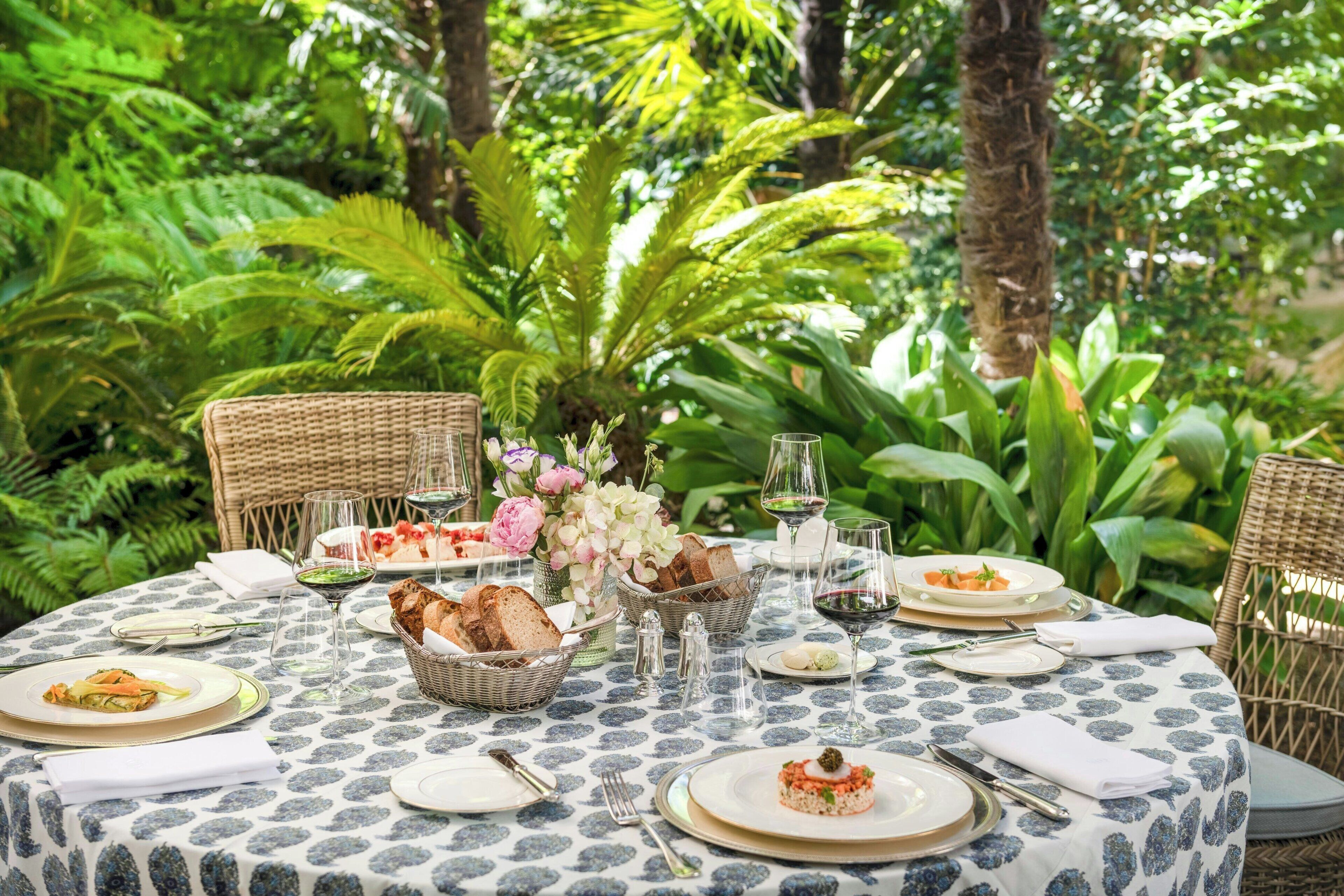 An outdoor dining area with a round table set for a meal, surrounded by lush greenery.