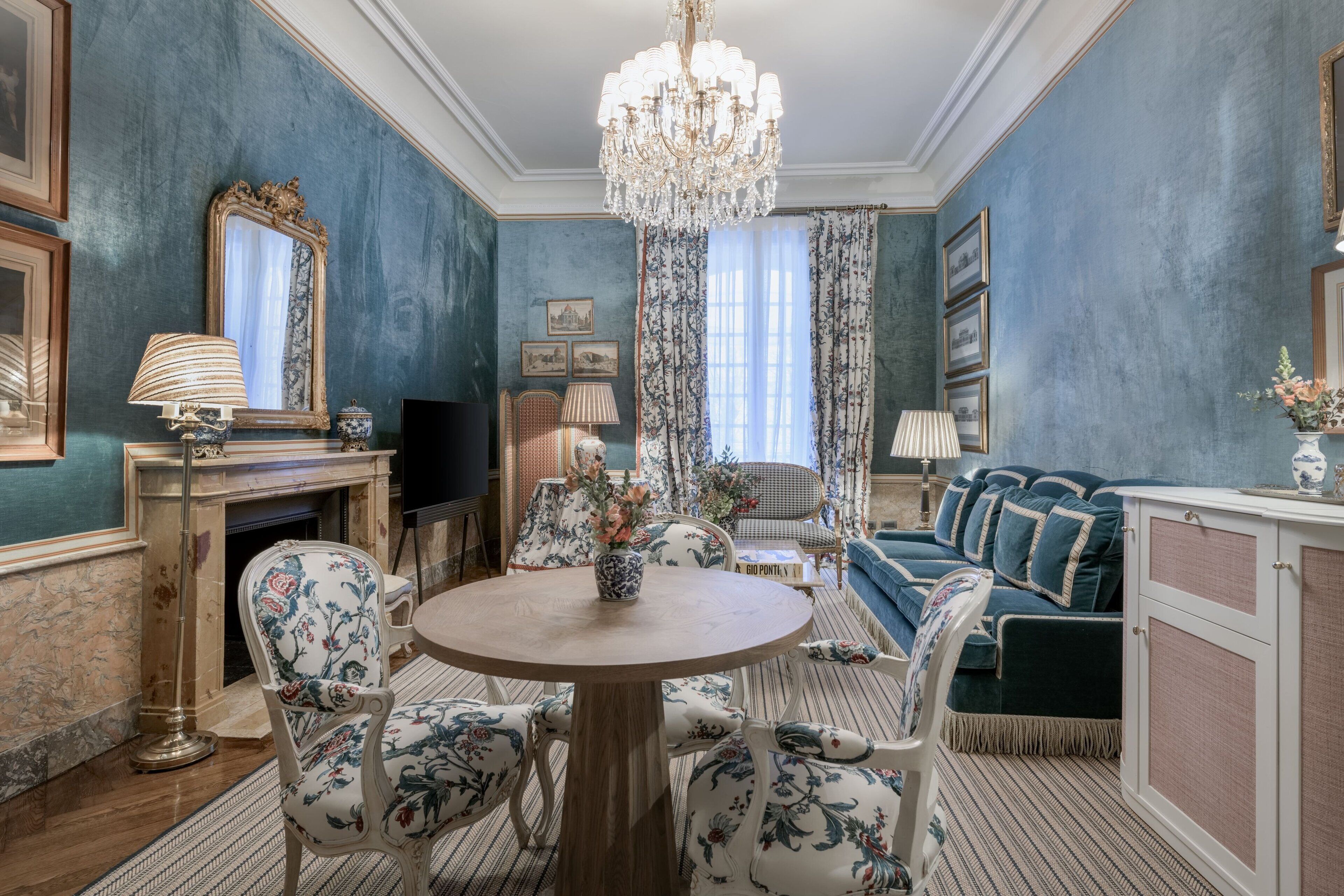 A room with a round wooden table surrounded by floral-patterned chairs, a blue sofa, a chandelier, and a fireplace.