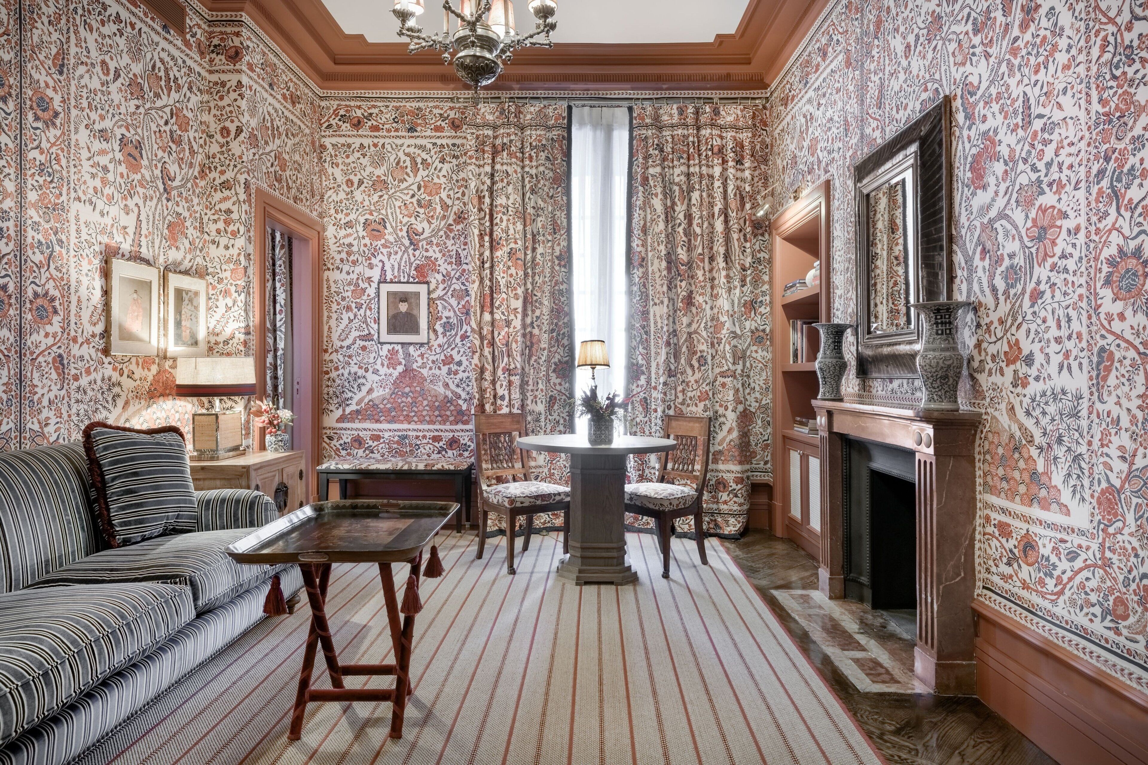A room with patterned wallpaper, a sofa, a table, chairs, a fireplace, and a chandelier.
