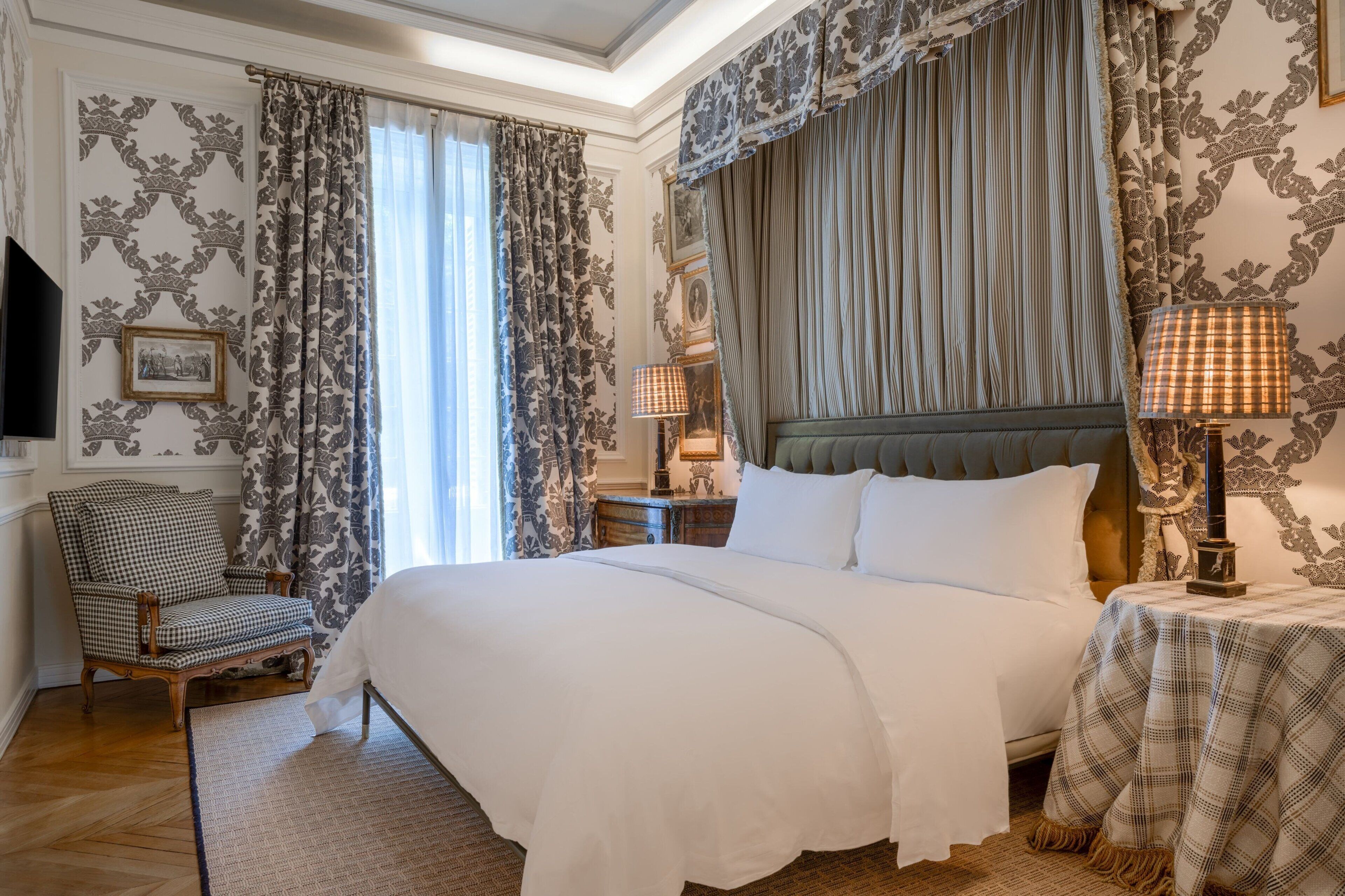 A hotel room with a large bed, a chair, a bedside table with a lamp, and patterned wallpaper.
