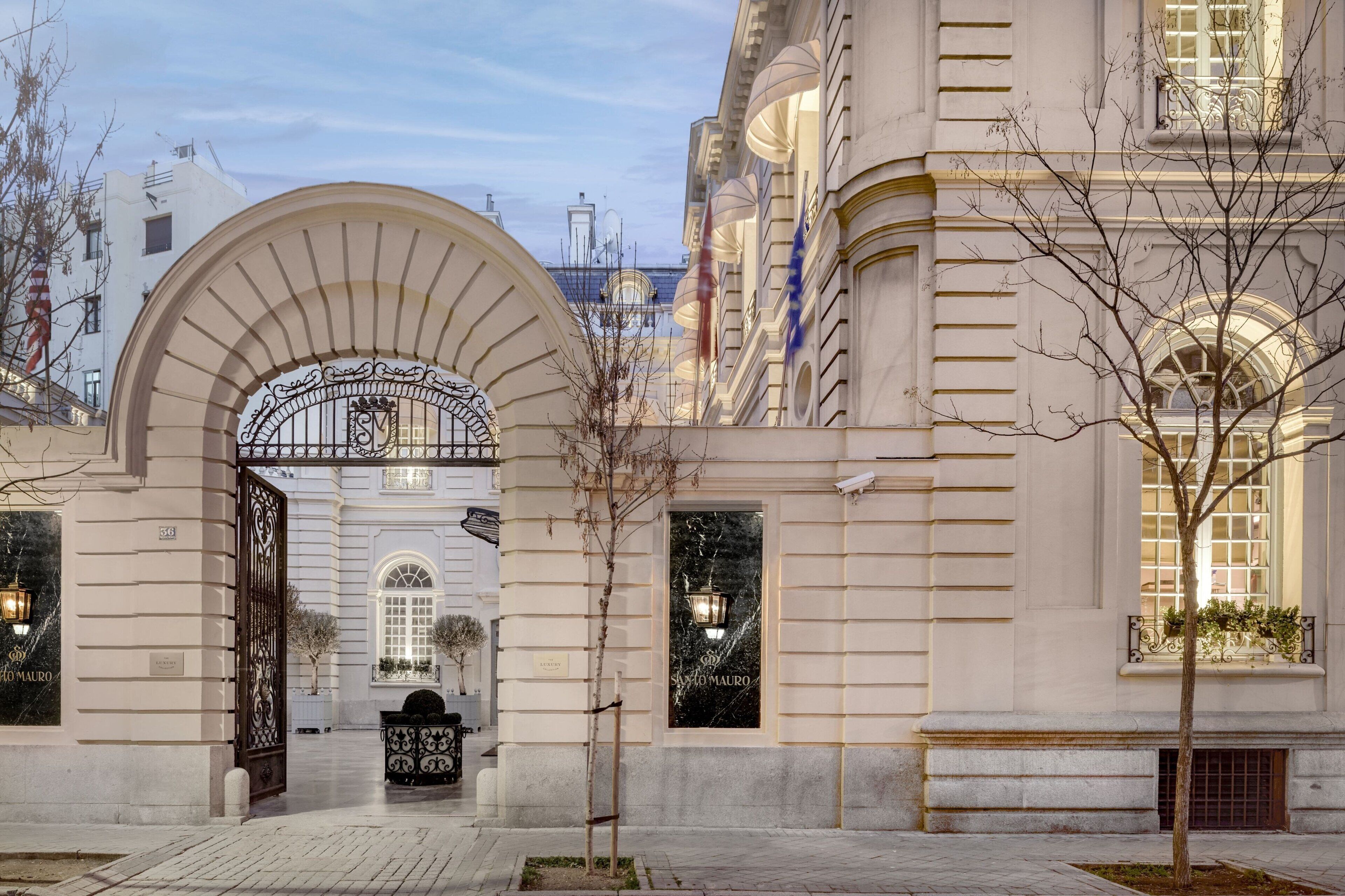 A grand entrance with an ornate metal gate, a well-maintained courtyard, and a building facade with large windows and decorative elements.