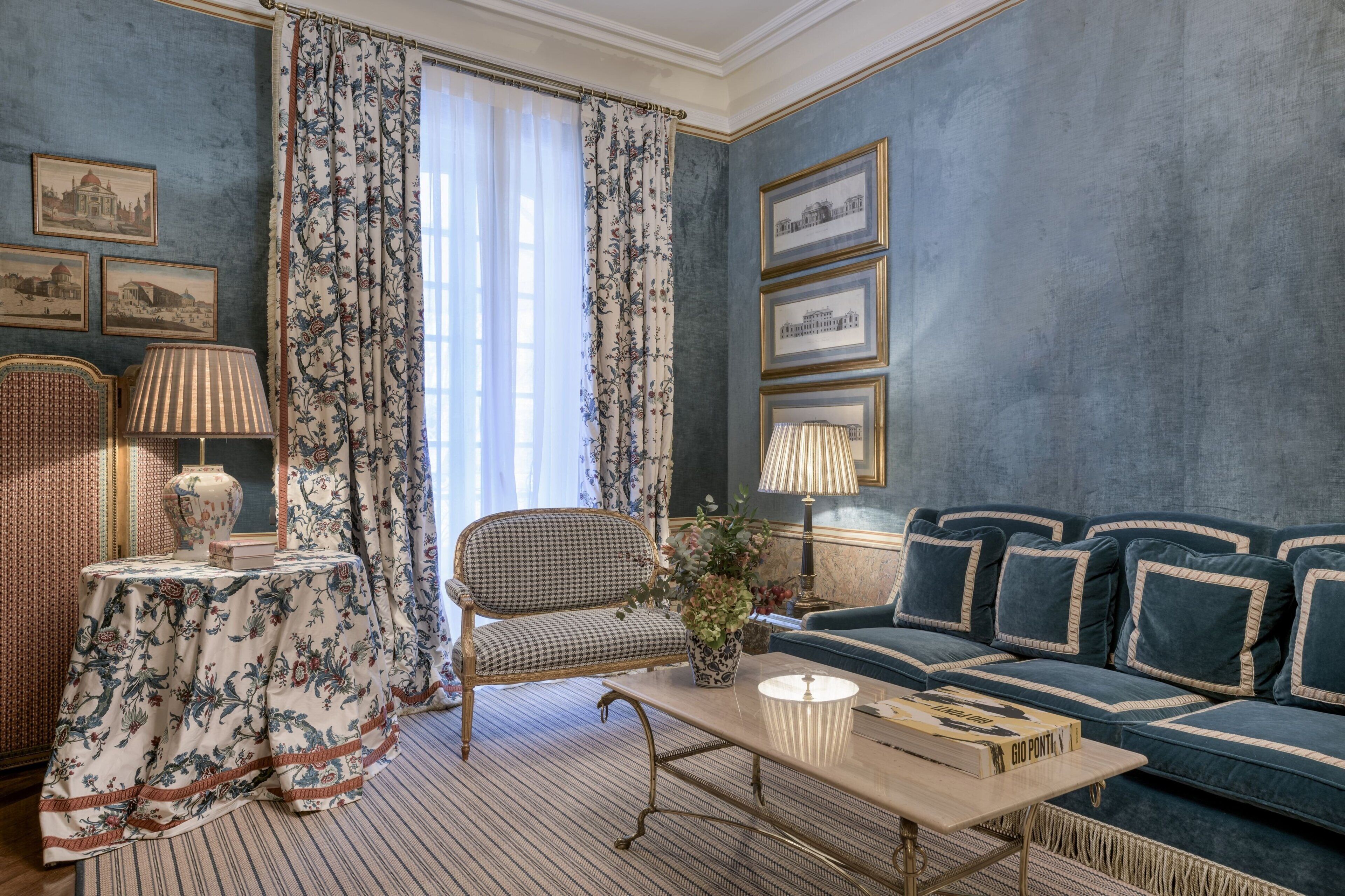 A room with a checkered sofa, a striped rug, a table with a lamp, and a window with floral curtains.