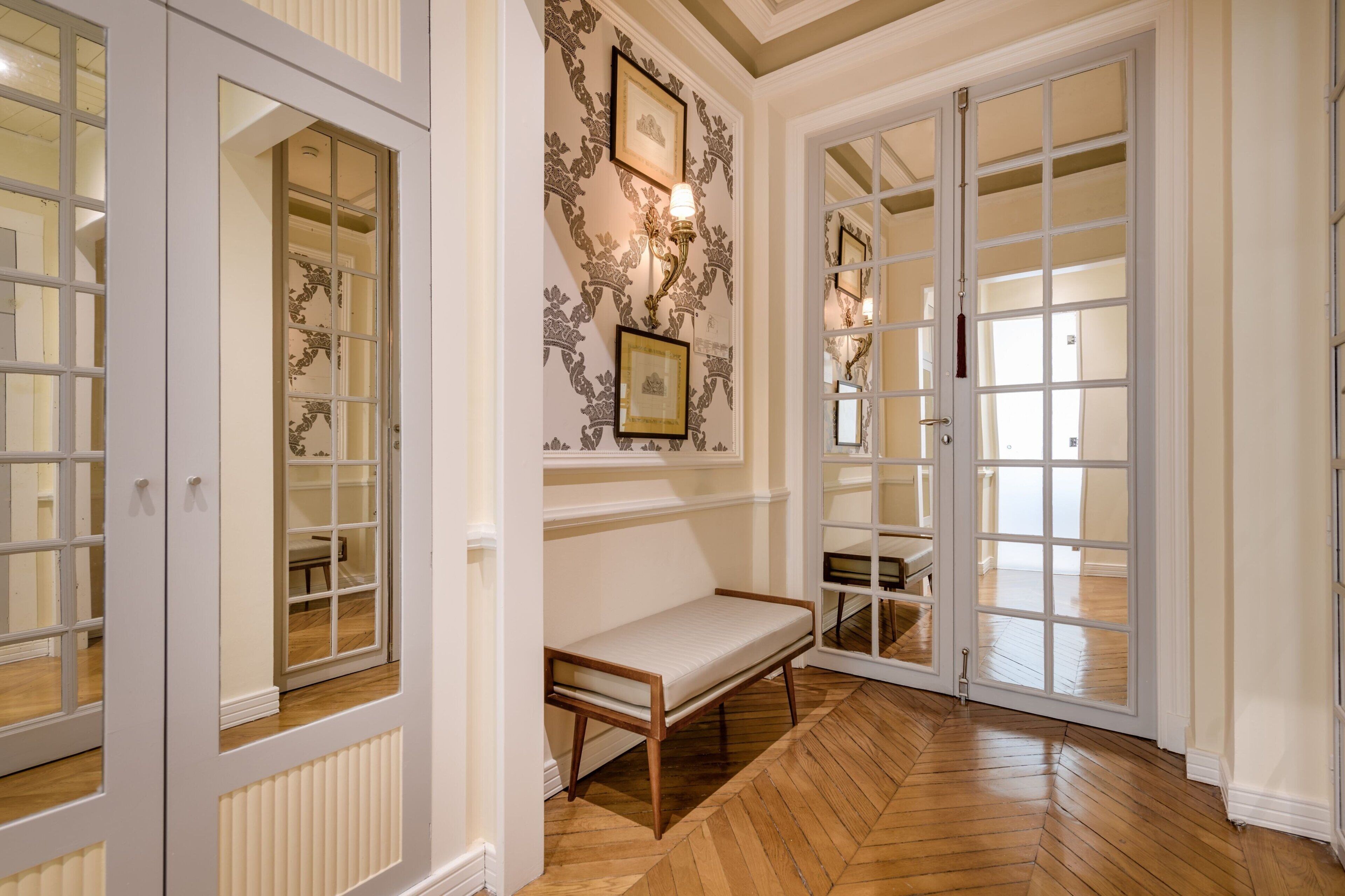 Un couloir avec un banc, une porte double dotée de panneaux de verre et un papier peint à motifs.