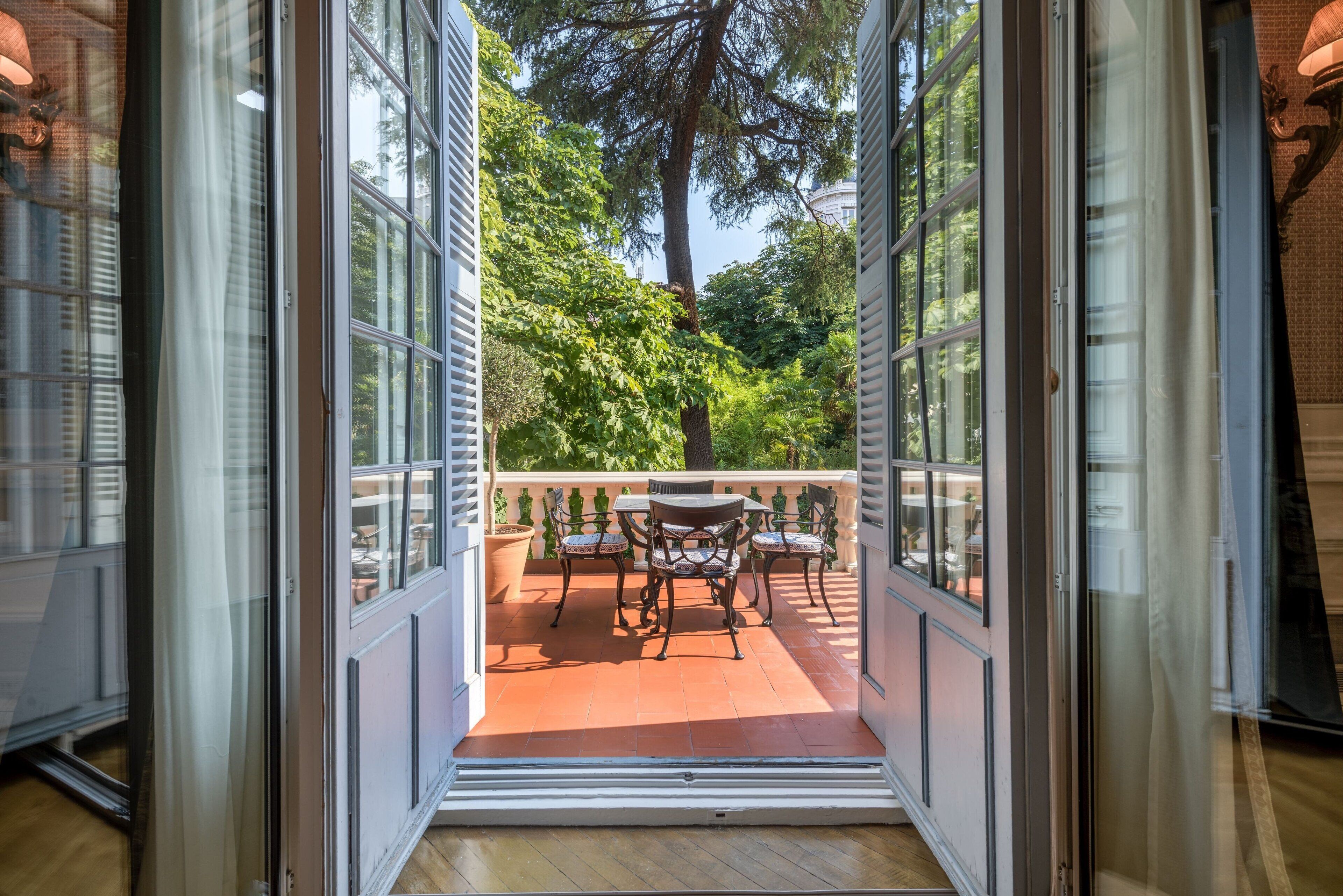 An open door leading to a patio with a table and chairs, surrounded by greenery and a view of trees.