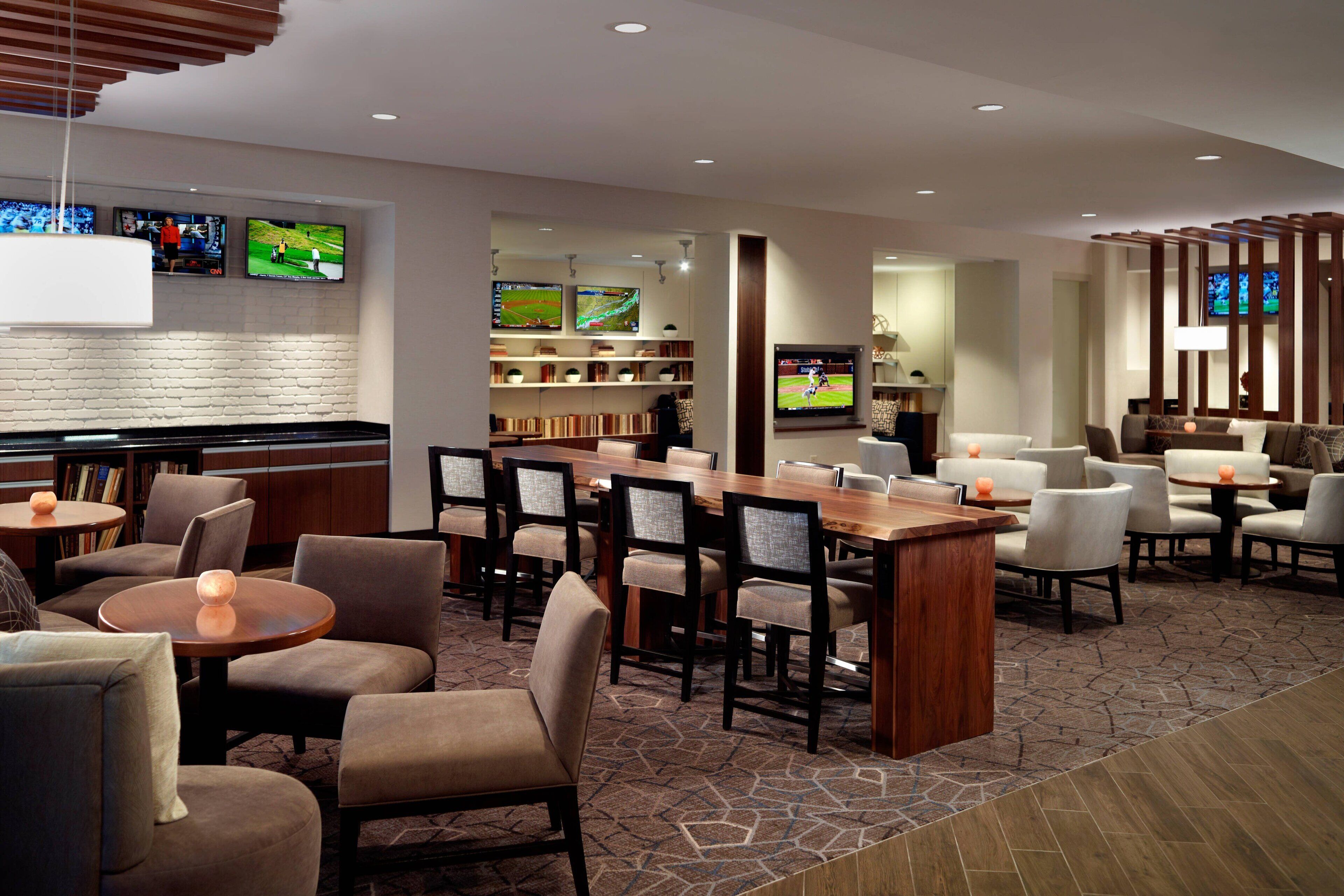 A modern hotel lobby with a bar area, multiple seating options, and a television screen displaying sports.