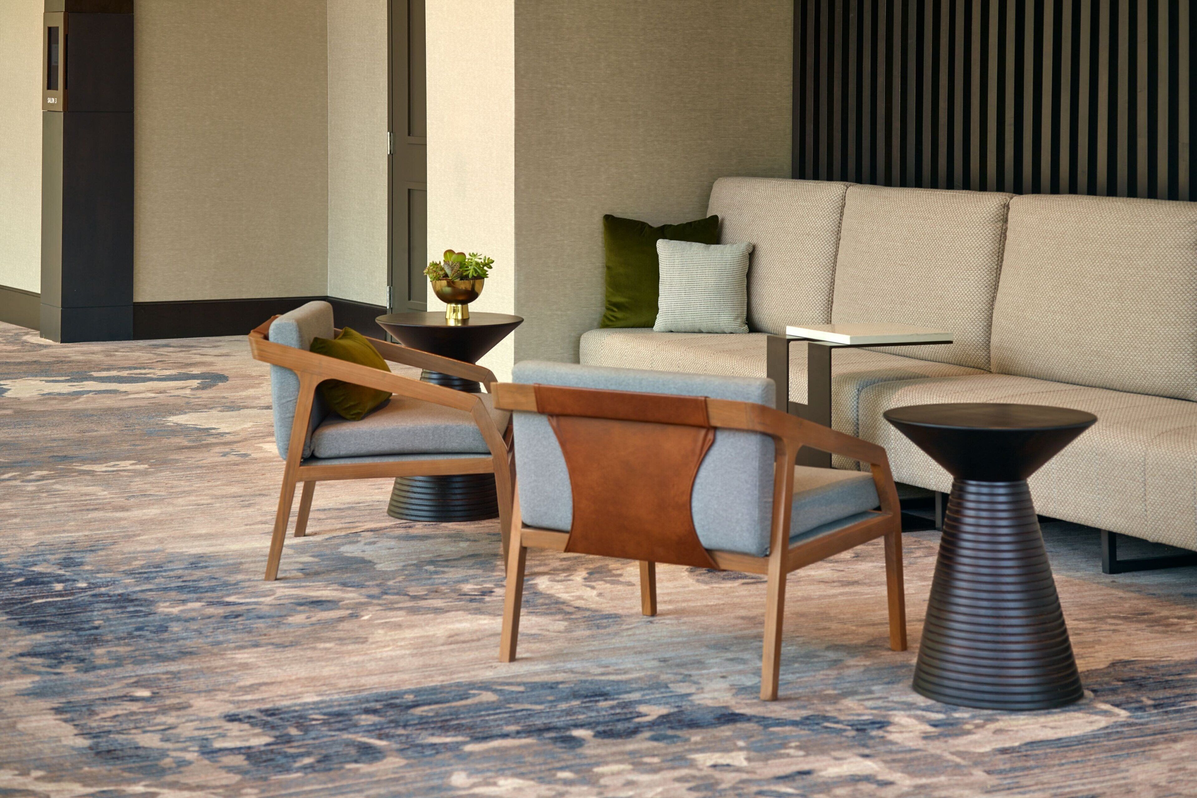 A modern lobby with a beige sofa, two wooden chairs with cushions, a small table, and a decorative plant.