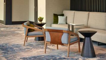 A modern lobby with a beige sofa, two wooden chairs with cushions, a small table, and a decorative plant.