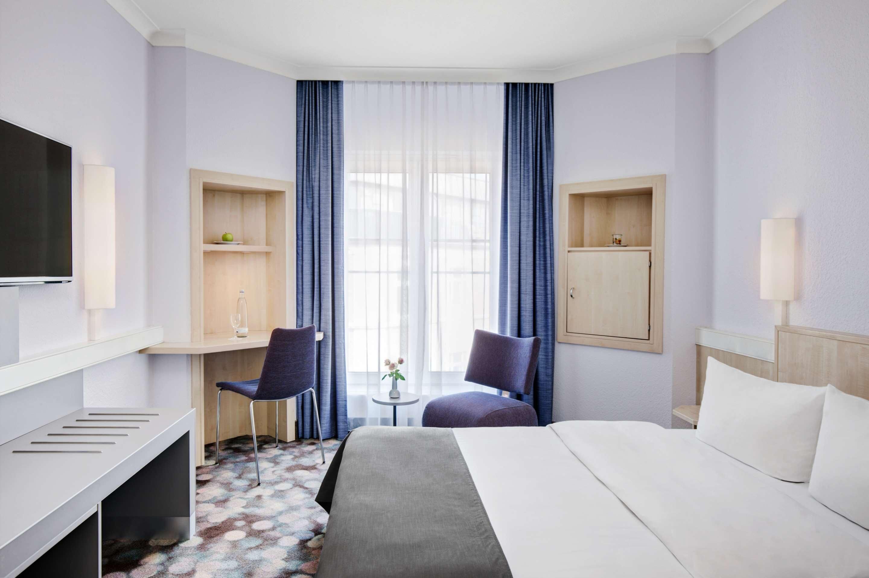 Habitación de hotel con una cama, dos sillas, un escritorio, un televisor y una mesita con flores.