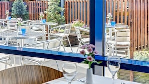 An outdoor dining area with round tables and chairs, a wooden fence, and potted plants. - IntercityHotel Hamburg-Altona (Hamburg)