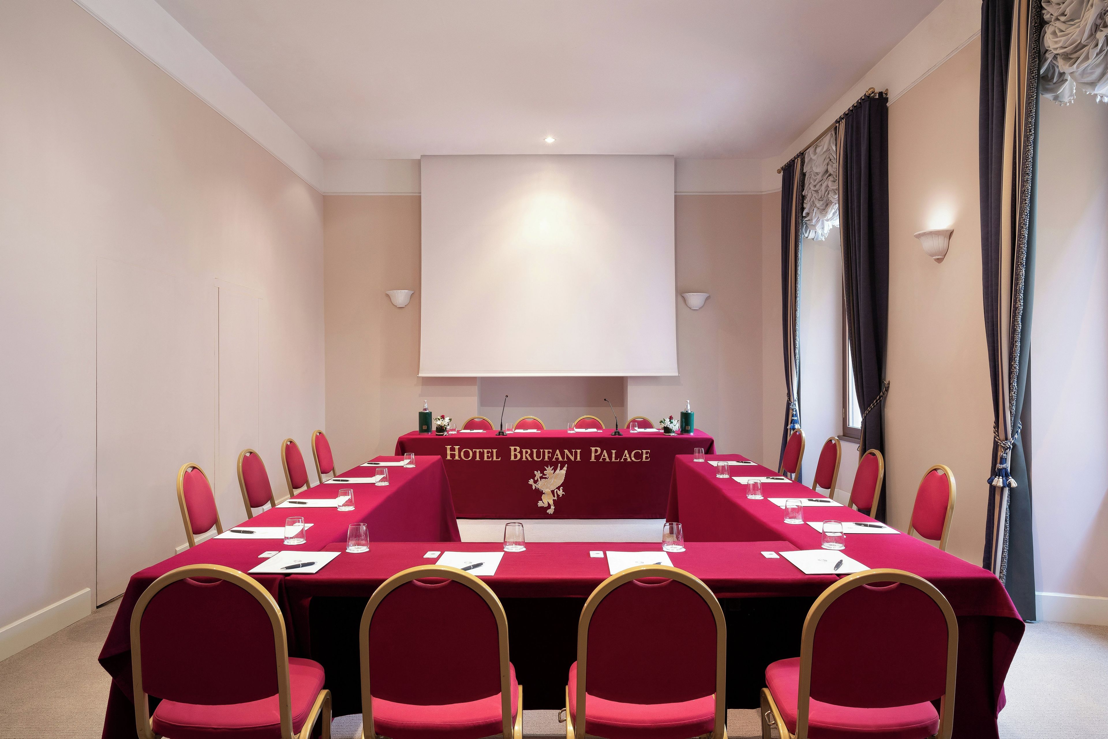 Ein Konferenzraum mit einem langen Tisch, roten Stühlen und einem Banner mit der Aufschrift 'Hotel Brufani Palace'.
