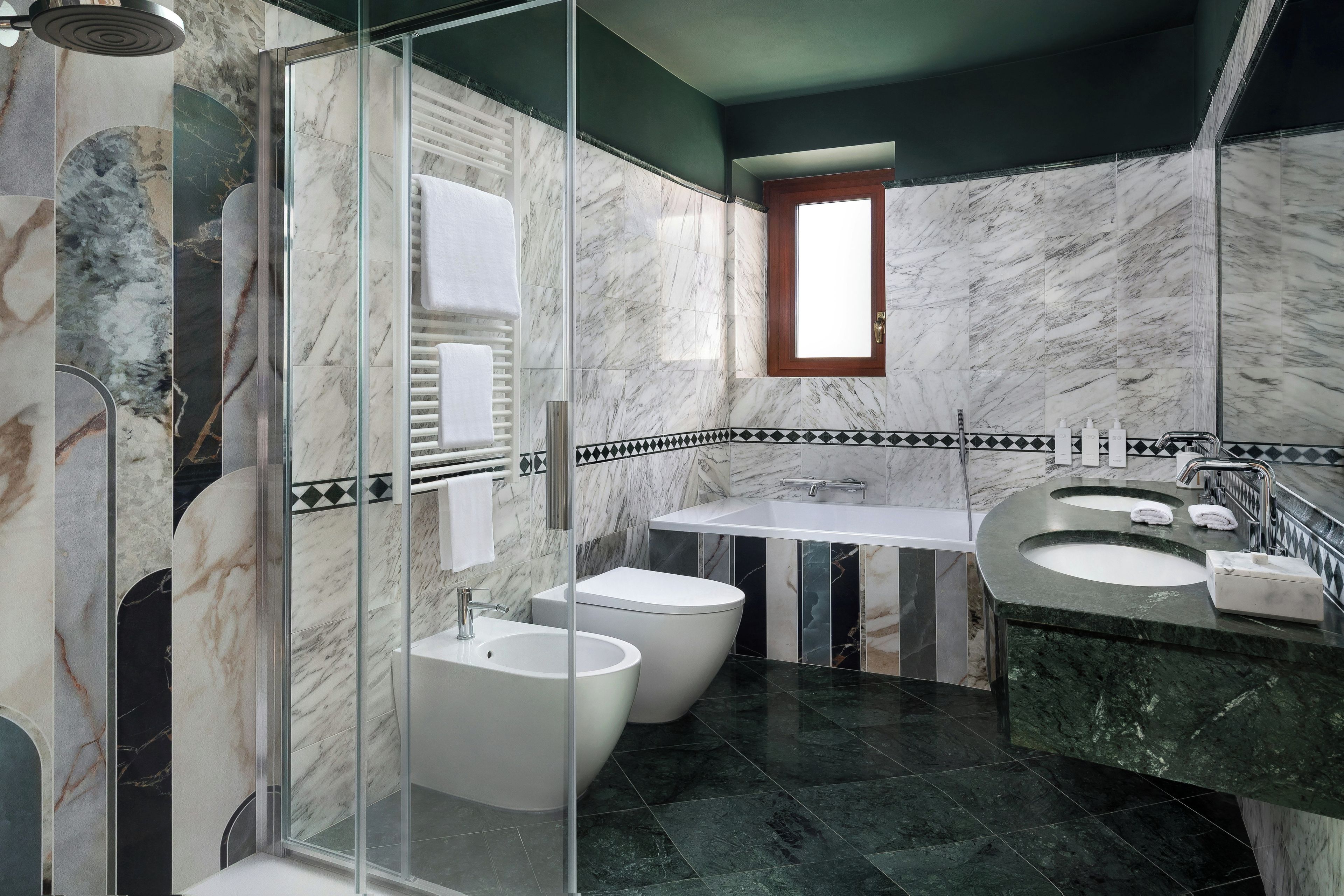 A modern bathroom with a glass-enclosed shower, two white bidets, a white sink, and a marble-tiled wall.