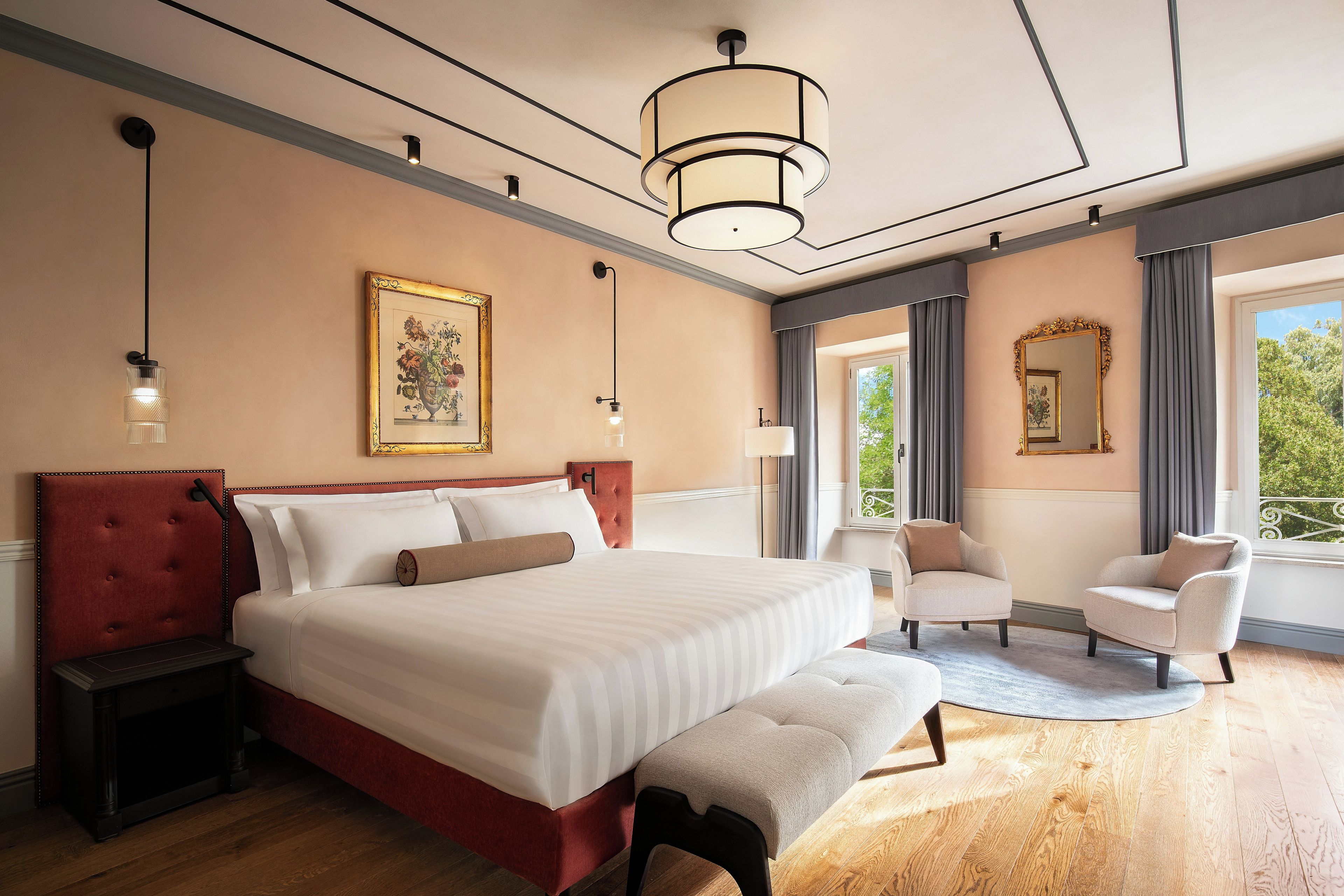 Habitación de hotel con una cama grande, mesitas de noche, un banco, una silla, un escritorio, una lámpara, un espejo y una ventana con vistas.