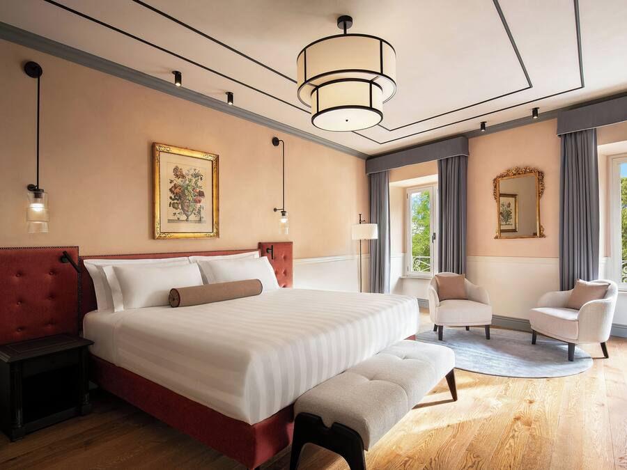 A hotel room with a large bed, bedside tables, a bench, a chair, a desk, a lamp, a mirror, and a window with a view.