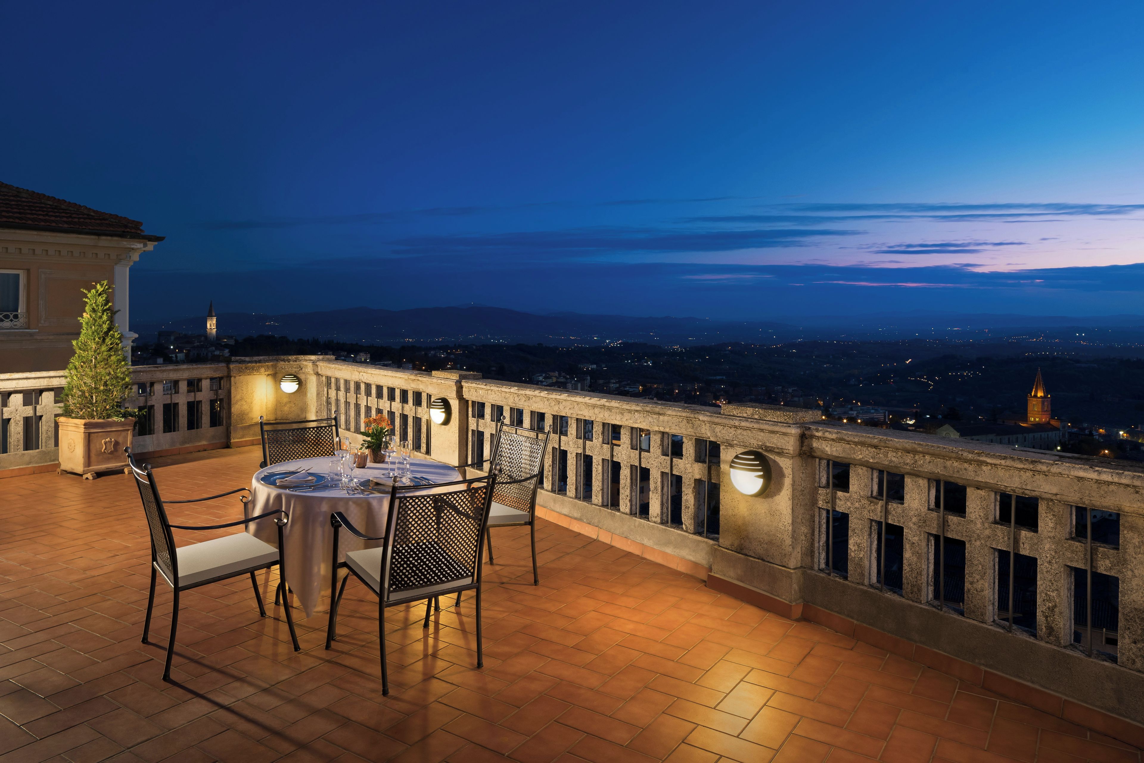 Una terraza con una mesa, sillas, barandilla y vista a un paisaje urbano al anochecer.