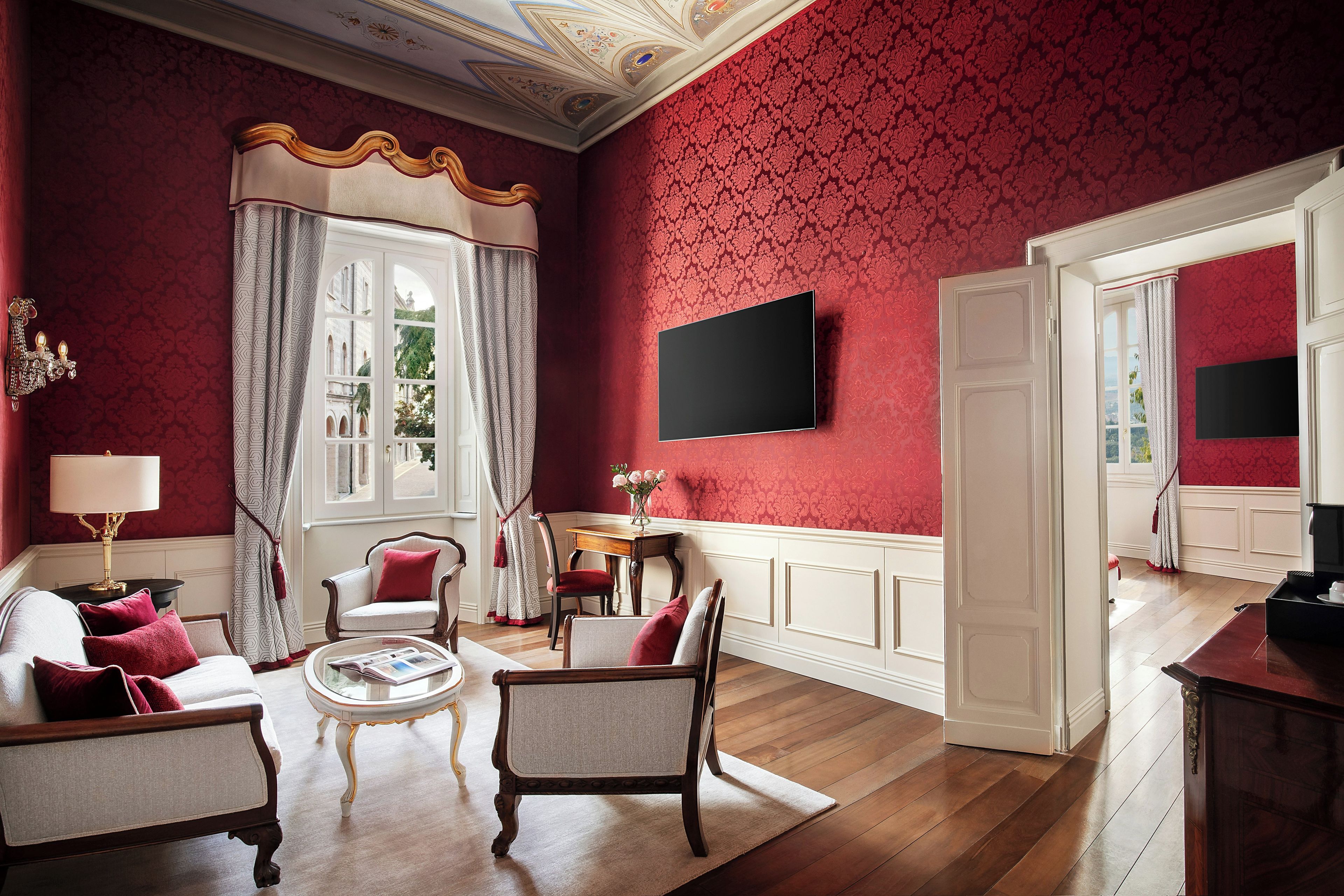 Une pièce avec un mur orné d’un motif rouge, un canapé blanc avec des coussins rouges, une chaise en bois, une petite table avec une lampe et un téléviseur à écran plat fixé au mur.