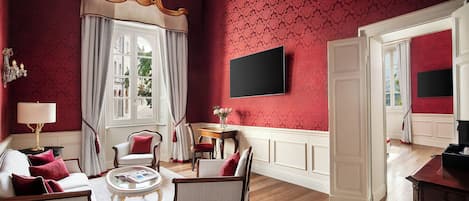 Une pièce avec un mur orné d’un motif rouge, un canapé blanc avec des coussins rouges, une chaise en bois, une petite table avec une lampe et un téléviseur à écran plat fixé au mur.