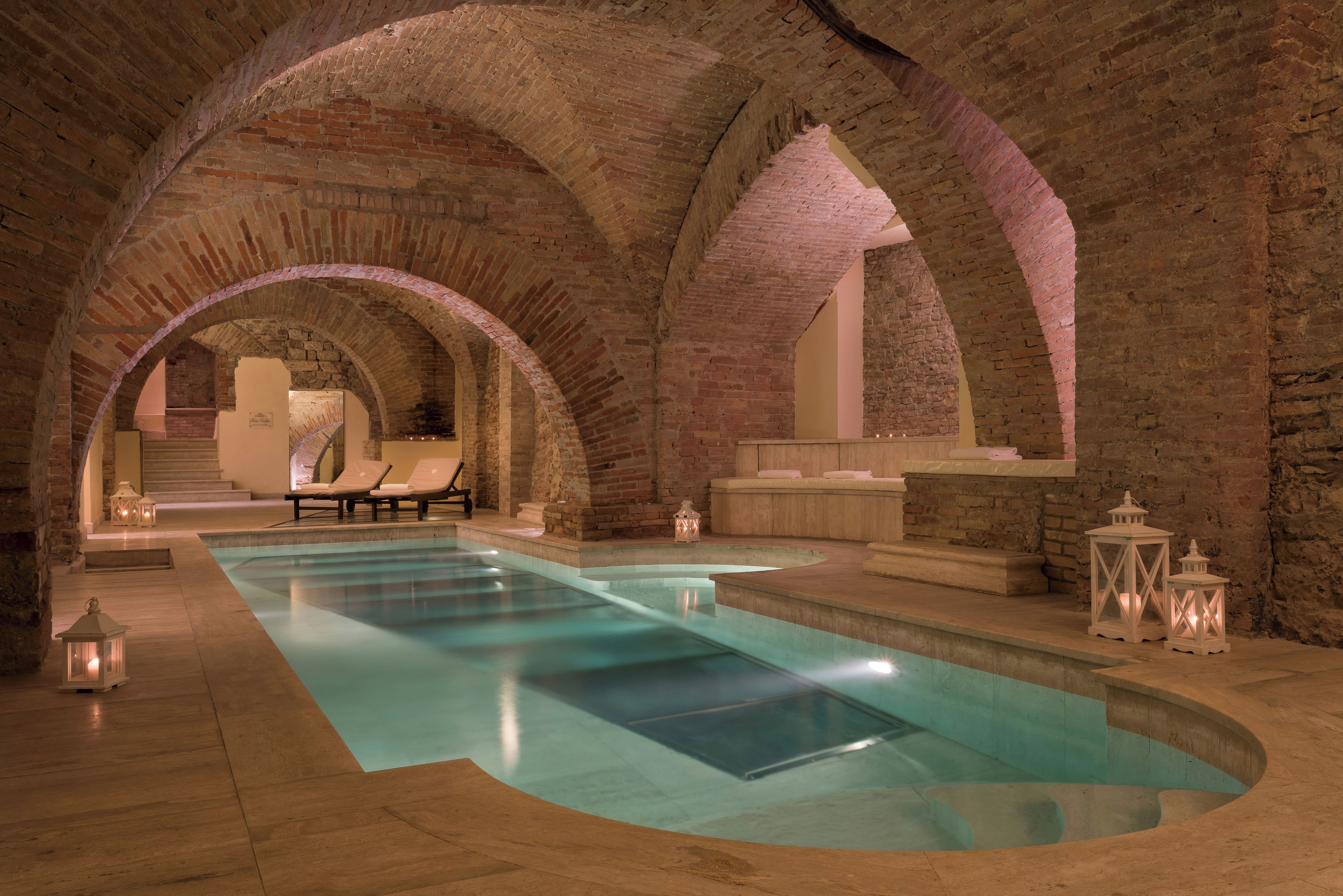 An indoor swimming pool with arched brick ceilings, lounge chairs, and decorative lanterns.